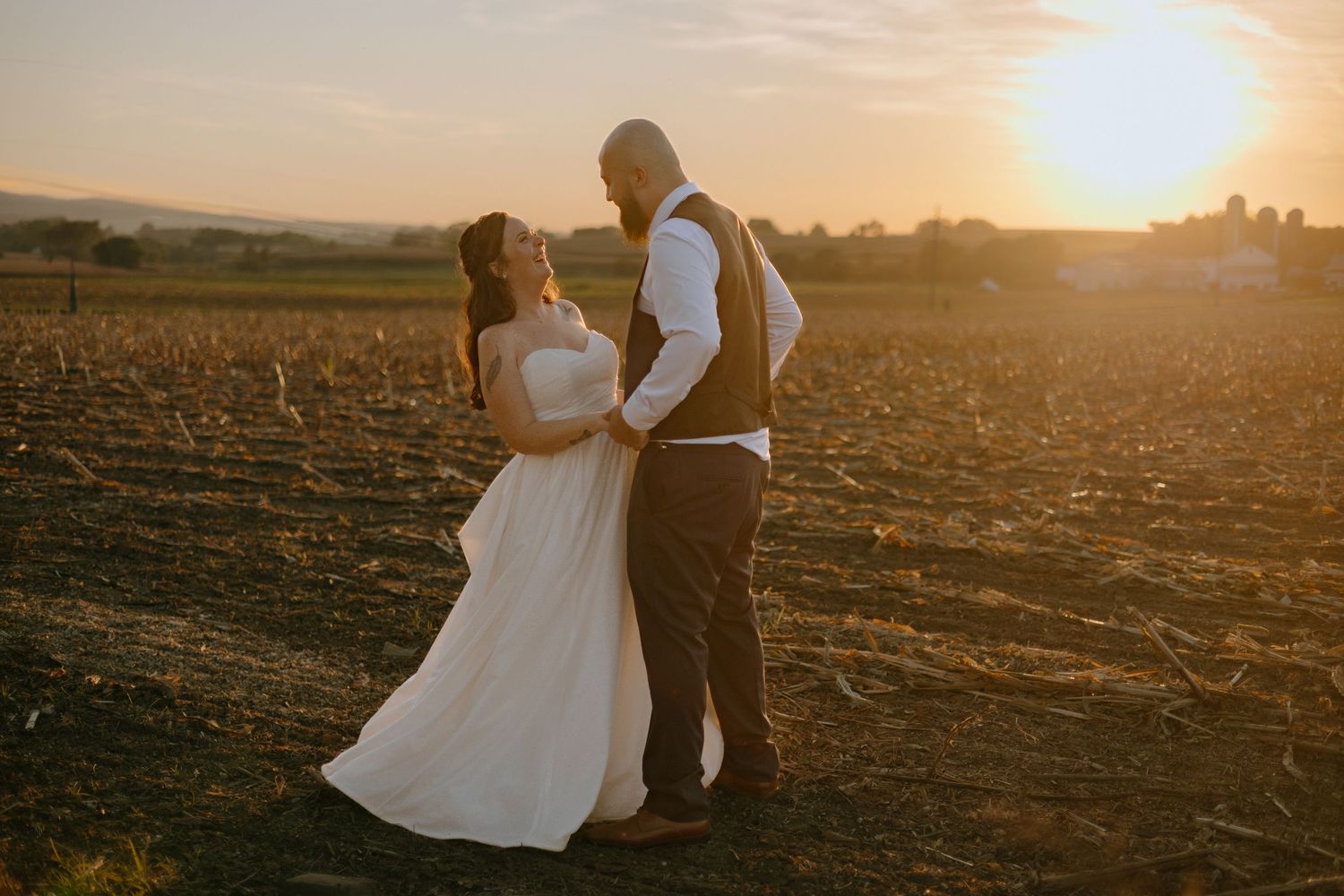 A couple in wedding attire embraces in a plowed field during golden hour sunset.