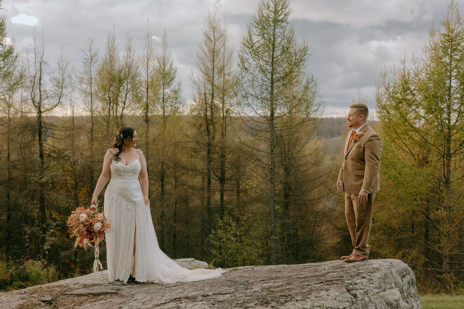 A couple in wedding attire shares an intimate moment on a rocky overlook surrounded by autumn trees.