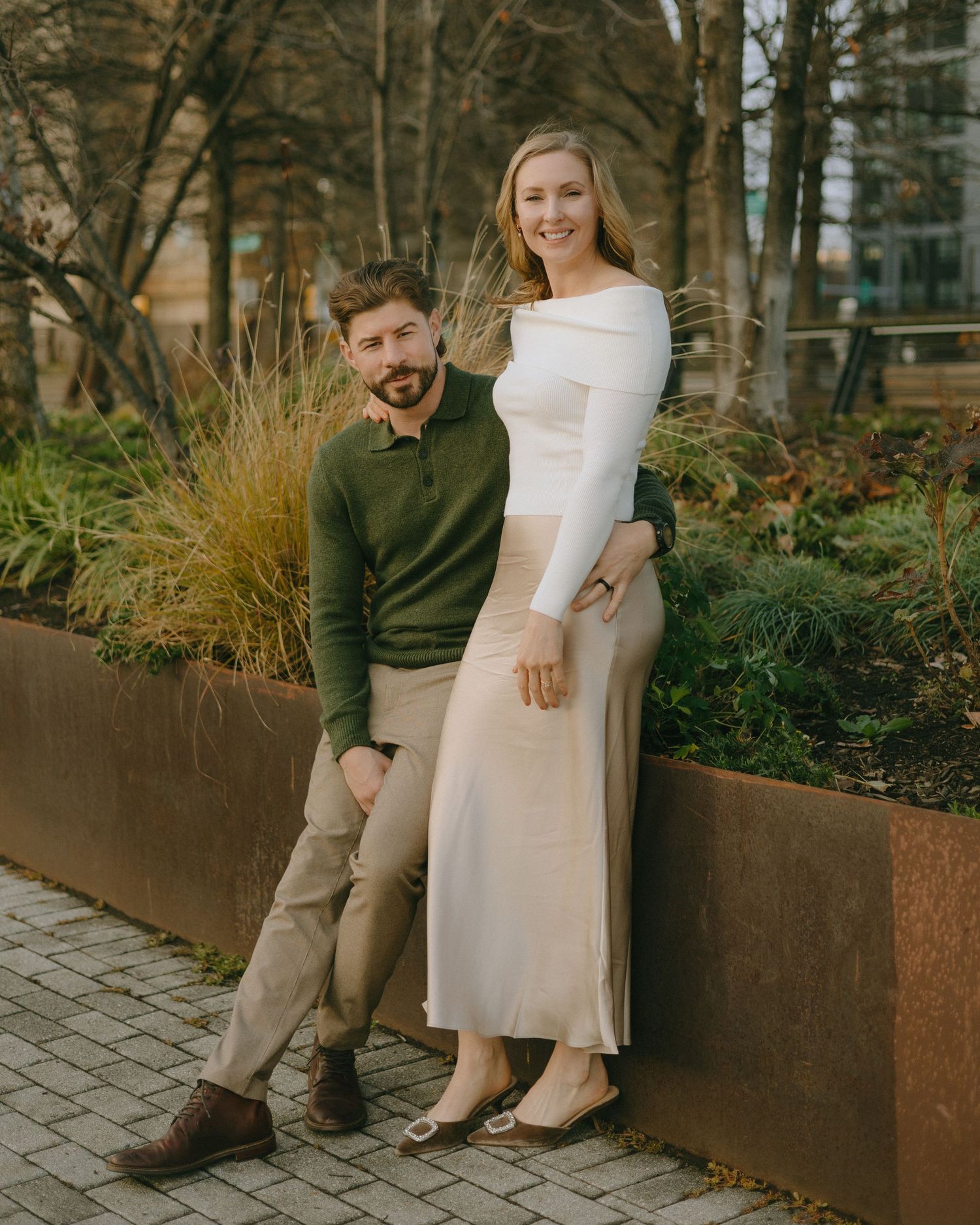 A couple poses casually on a concrete ledge in an urban garden setting with decorative grasses in the background.
