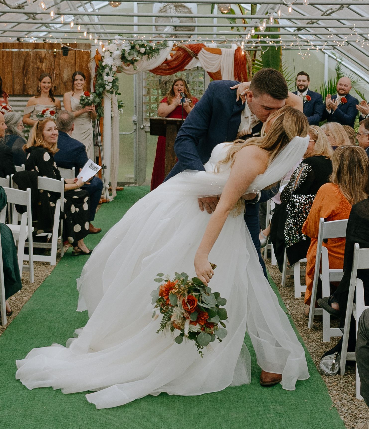 A wedding couple shares a playful dip pose during their outdoor ceremony on artificial turf with guests watching.