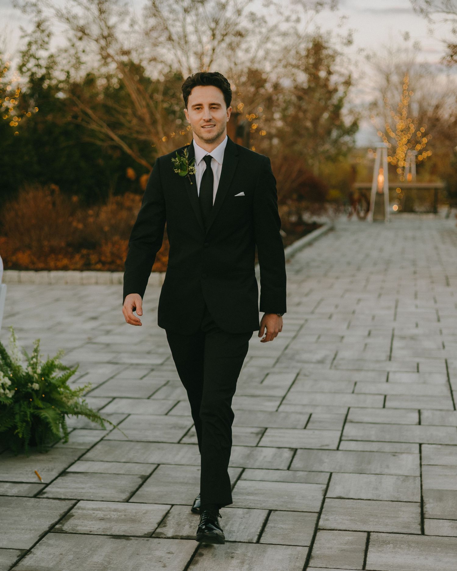 Groom in classic black suit walks on stone pathway with fall trees and landscaping.