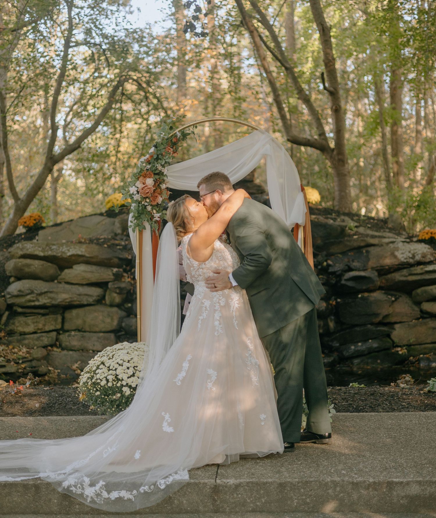 A couple shares romantic moments in a garden setting with stone walls and autumn foliage.