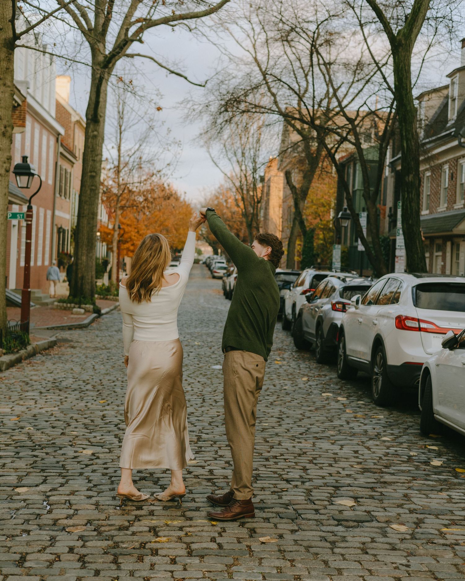 Two people stroll together on a historic cobblestone street lined with autumn trees and parked cars.