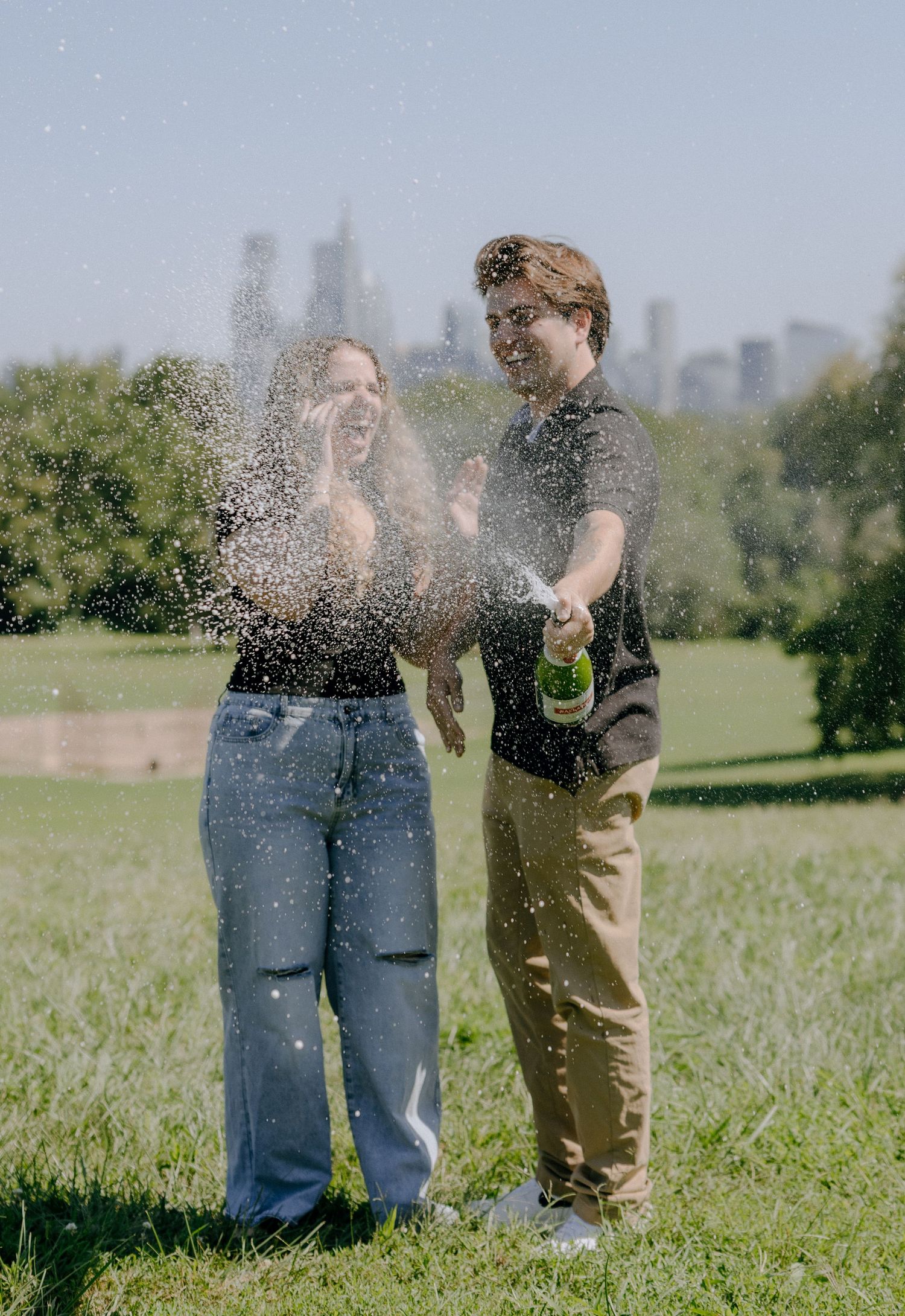 Champagne spray creates a festive moment between two people celebrating in a park.