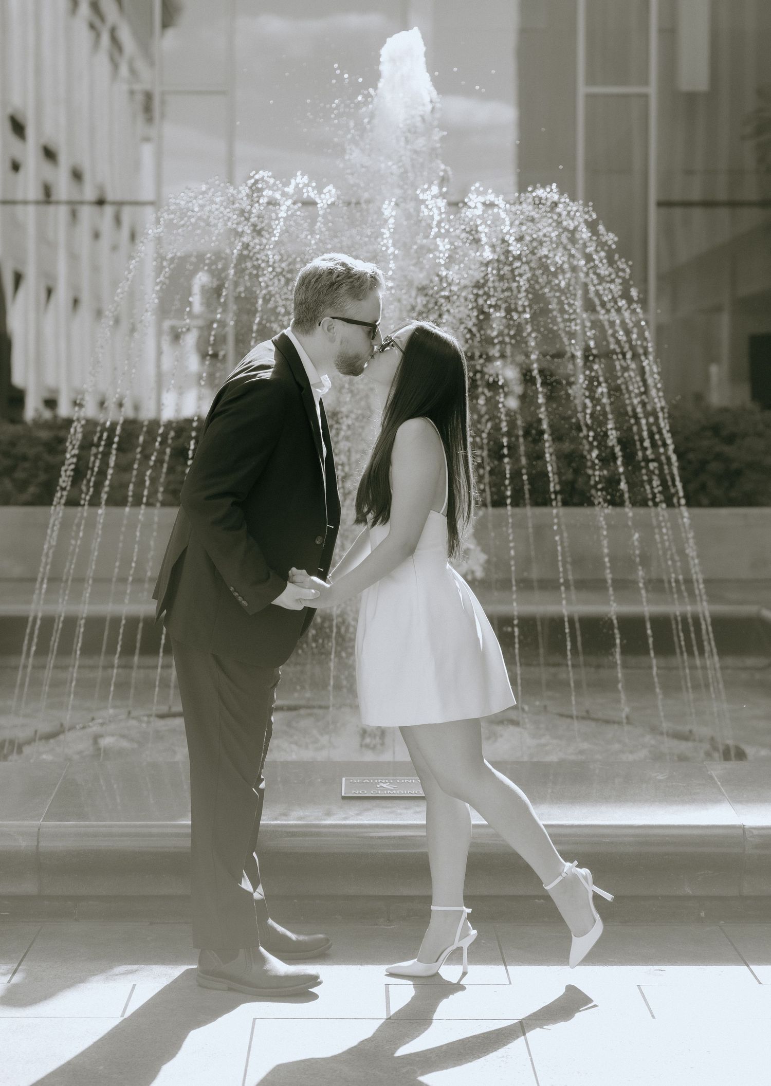 Black and white photo of couple dancing in formal attire near a water fountain.