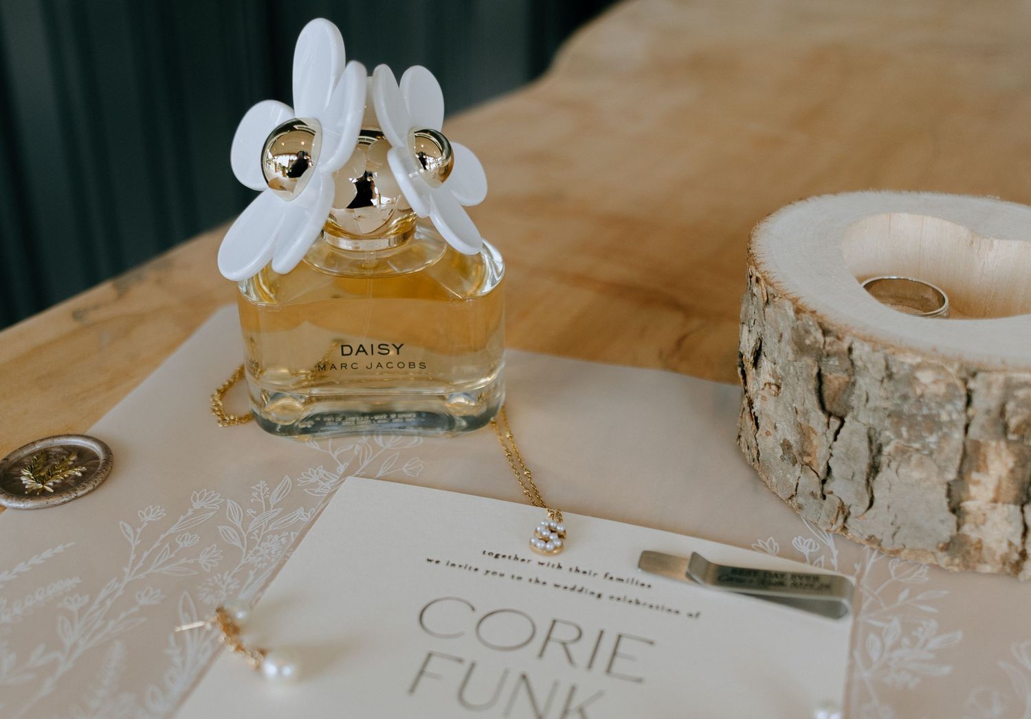 Perfume bottle with white floral decoration next to wedding invitation and birch candle holder on table.