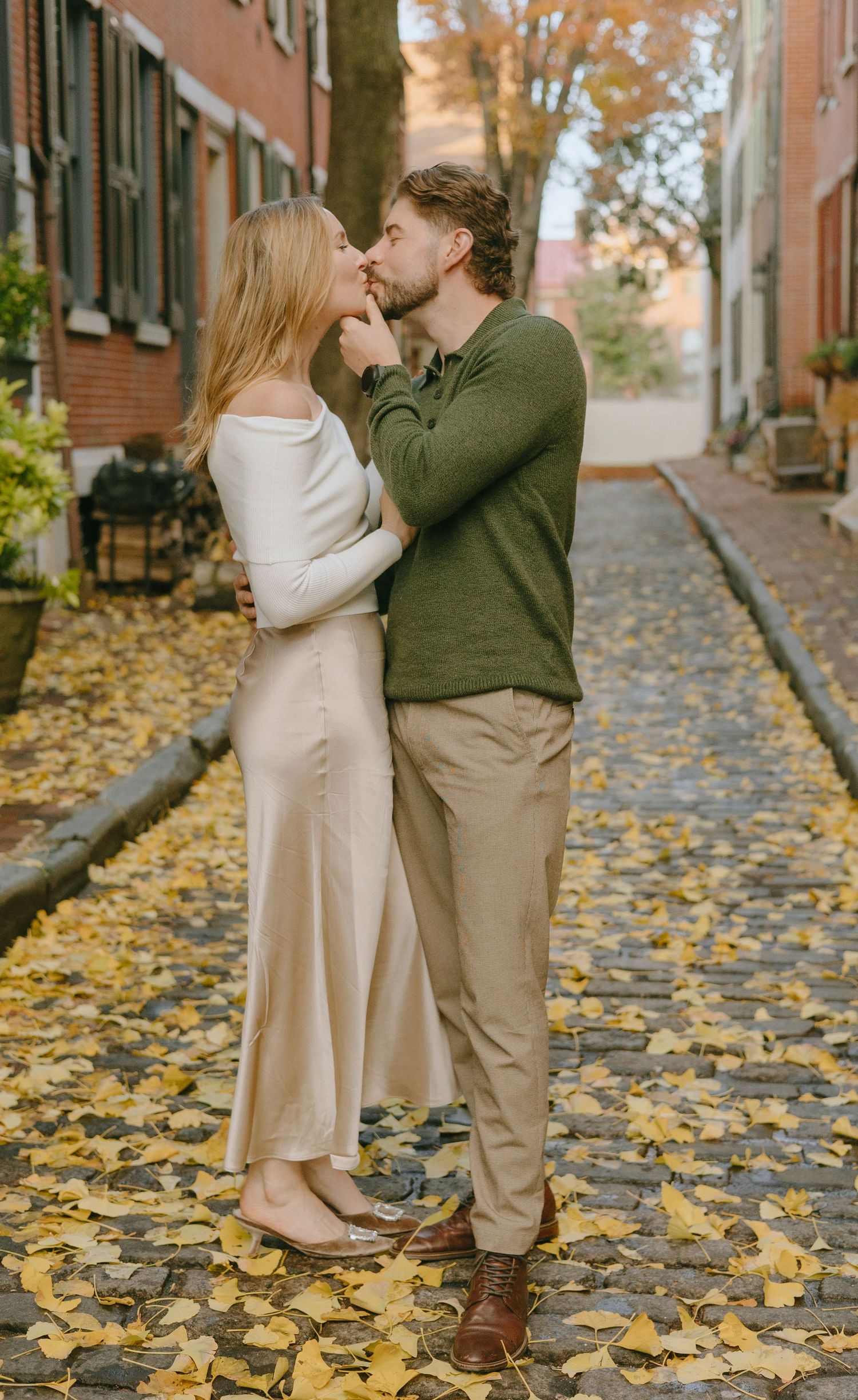 A couple shares a romantic moment on a cobblestone street covered in golden autumn leaves.