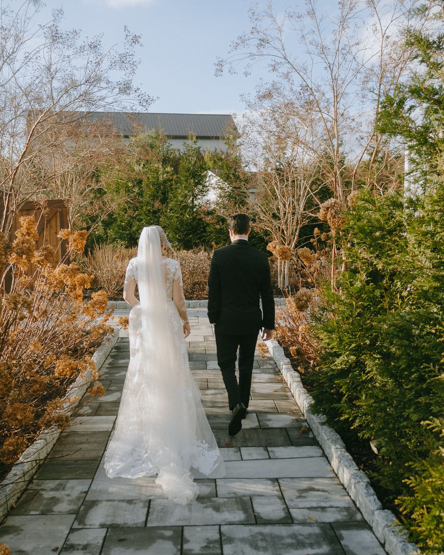 Bride in flowing white gown and groom in black suit walk along a stone path surrounded by autumn foliage.