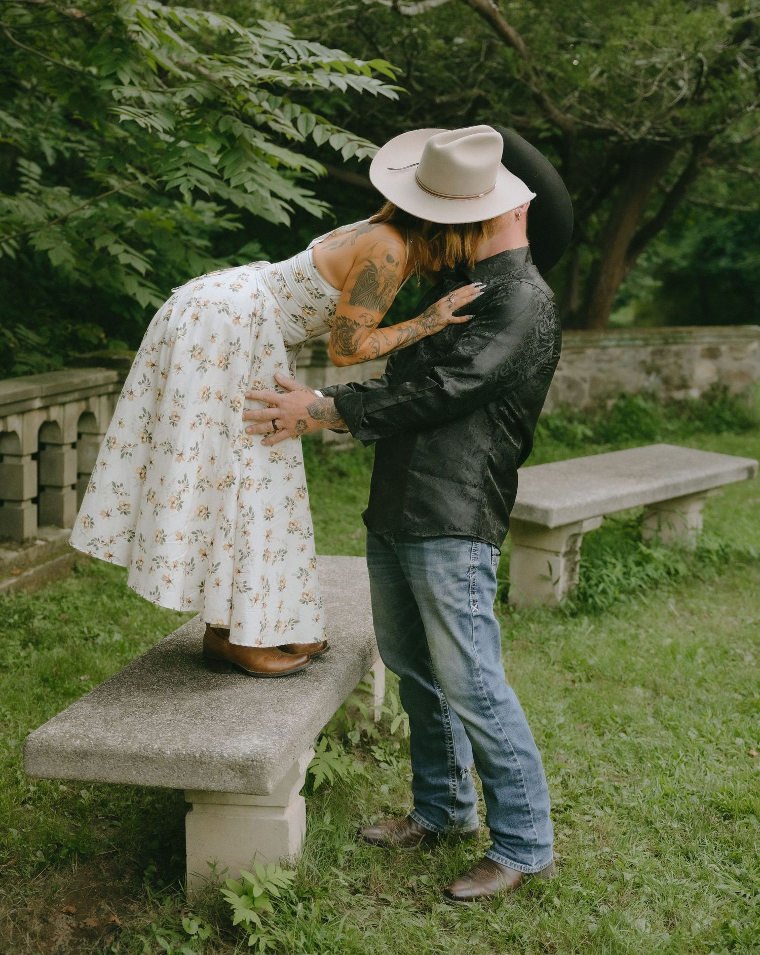 Couple in western wear plays and poses near stone benches in garden setting.