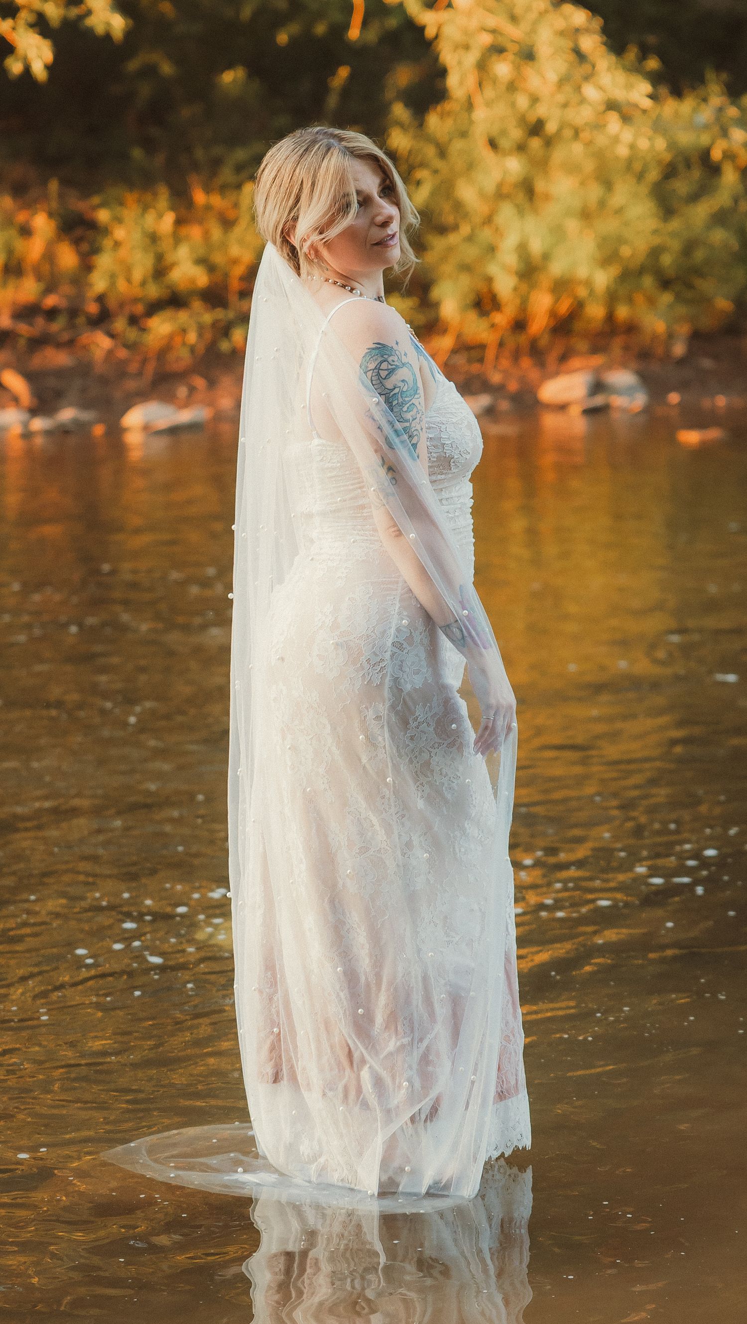 A bride's white lace dress becomes translucent as she stands in golden-lit river water.