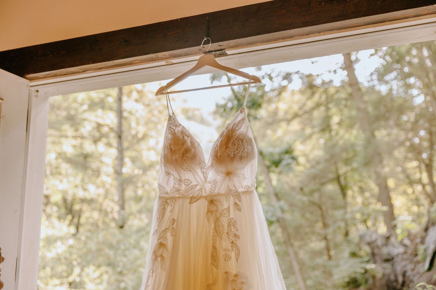 An elegant white wedding gown hangs in a sun-filled window gallery.