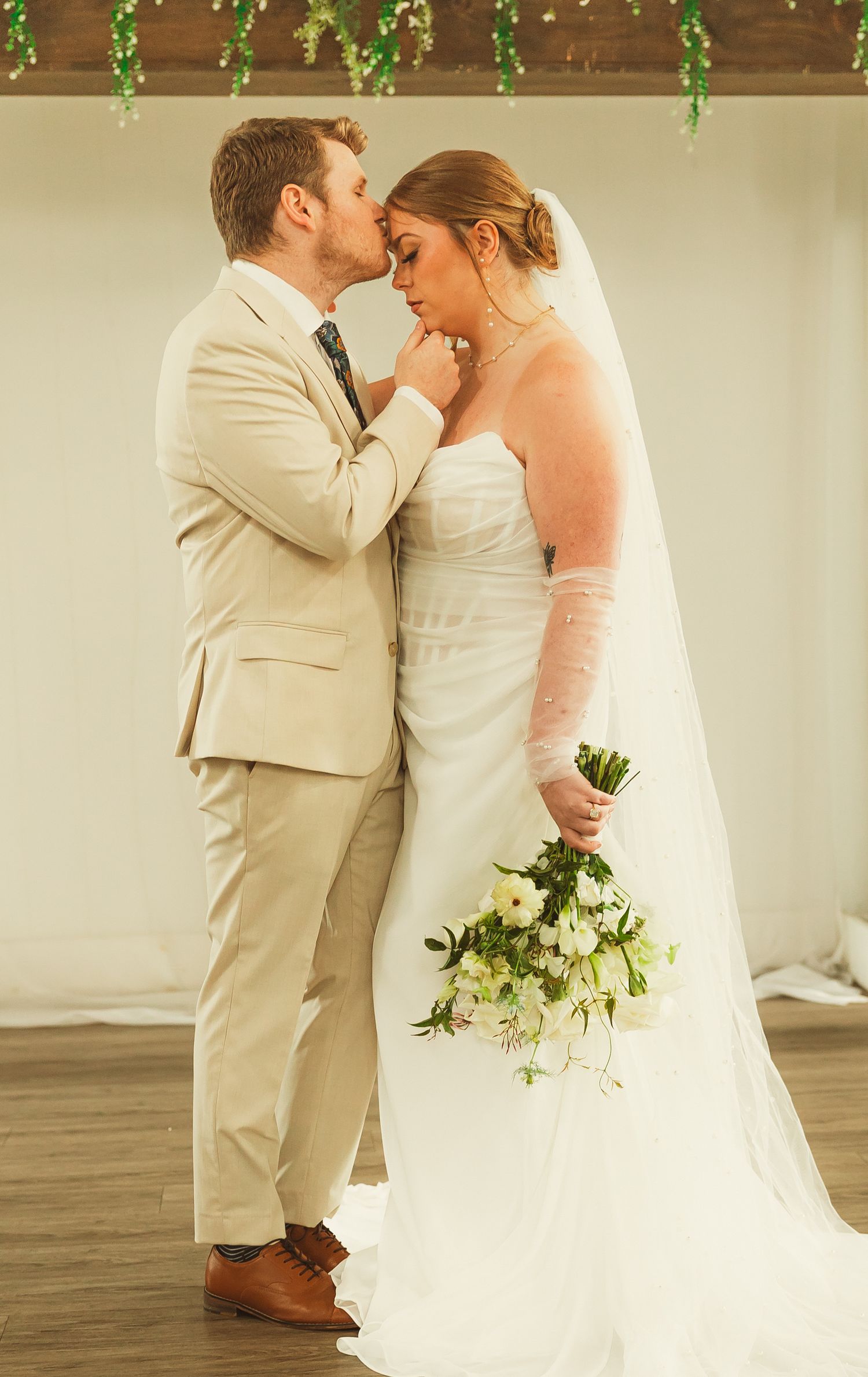 Romantic wedding couple embraces under hanging greenery decorations.