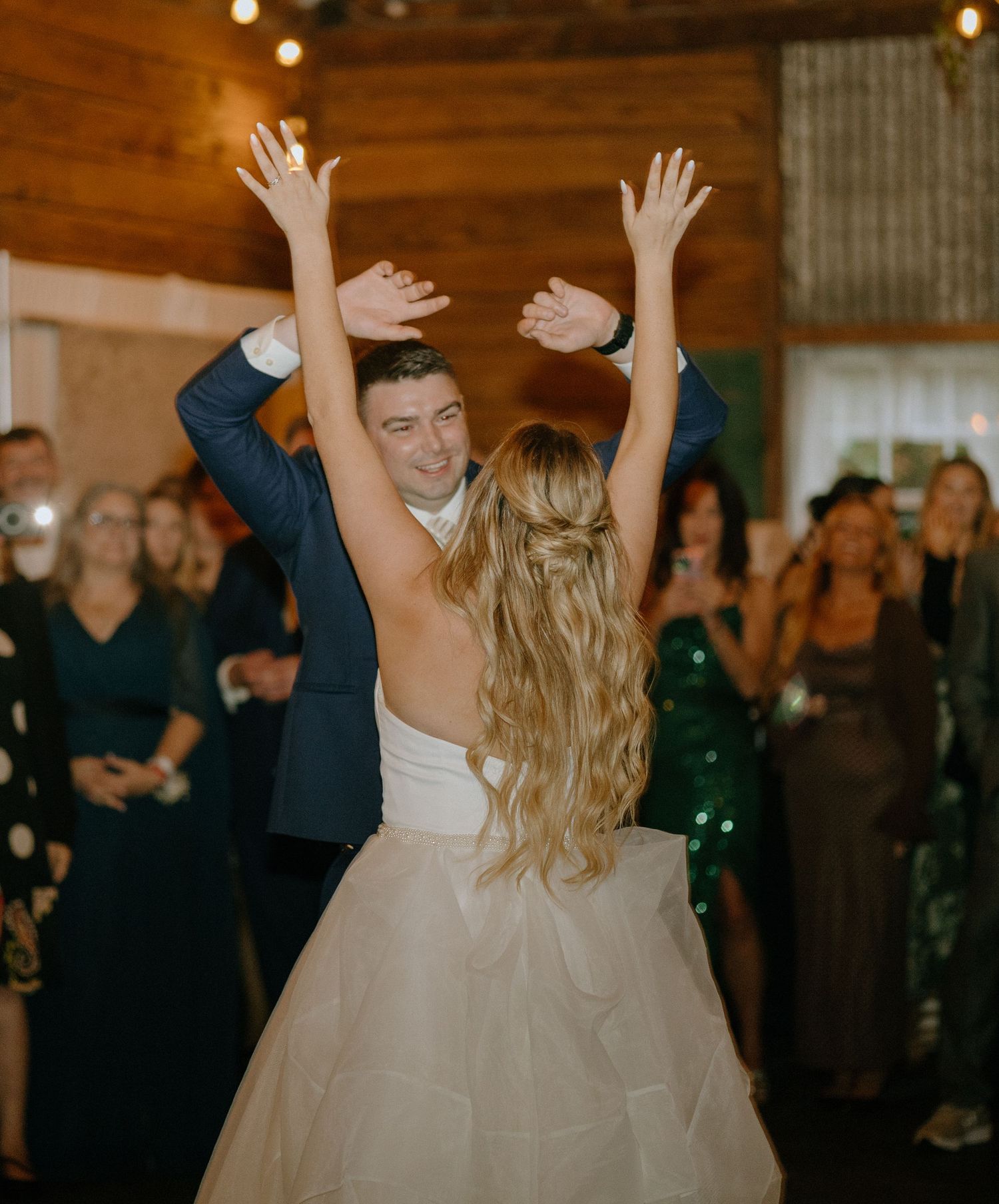 Wedding guests dance and celebrate on a dimly lit dance floor with string lights overhead during a reception.