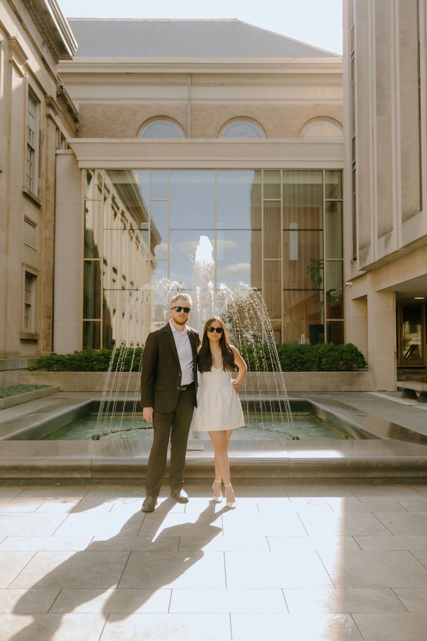 Couple posing near a fountain outside an elegant building with classical architecture.