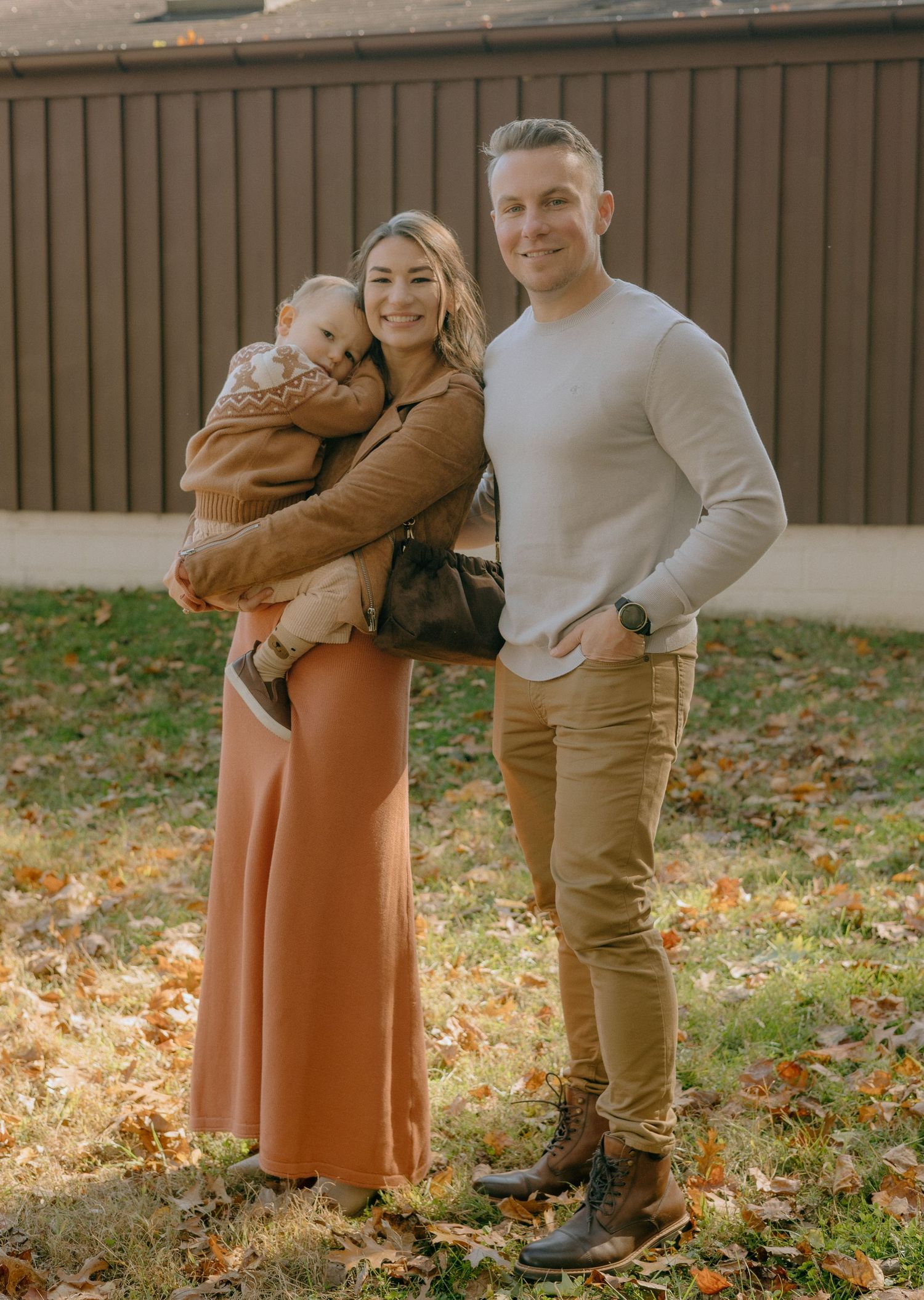 A couple in casual fall attire shares tender moments on fallen leaves in a grassy outdoor setting.