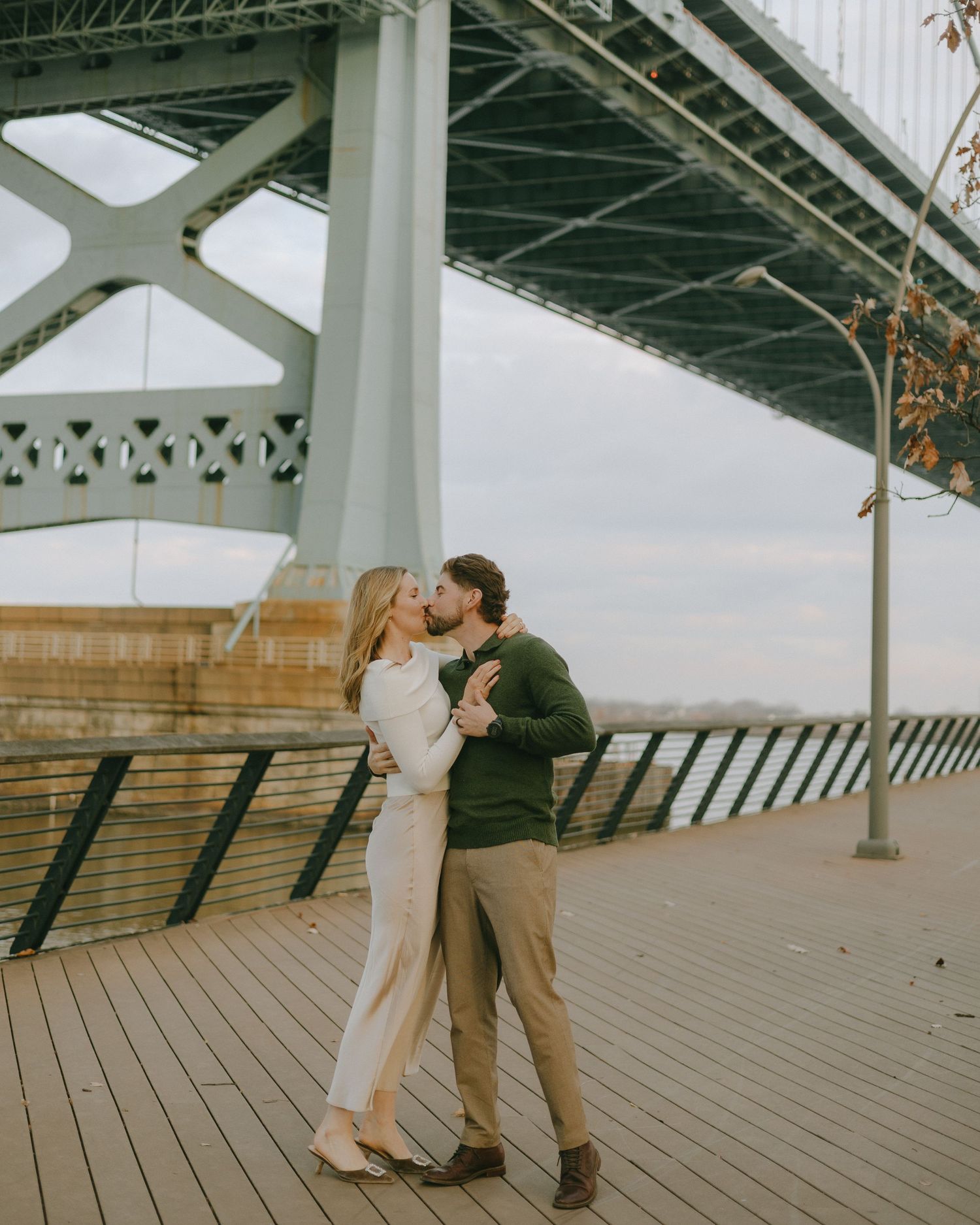 A romantic moment shared between a couple embracing on a wooden pier beneath a large bridge structure.