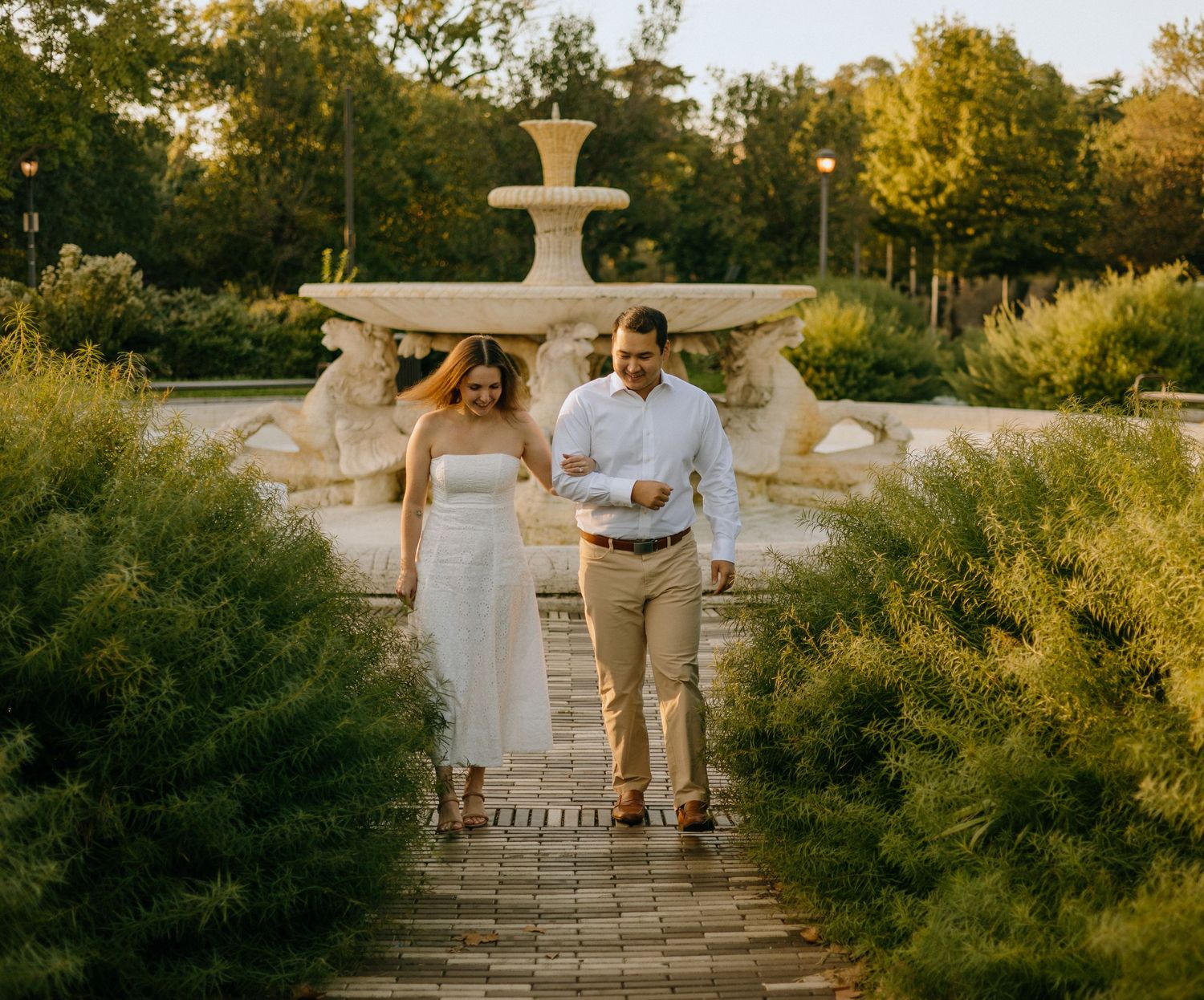 A couple in wedding attire walks up stone steps surrounded by lush greenery in a romantic garden setting at sunset.