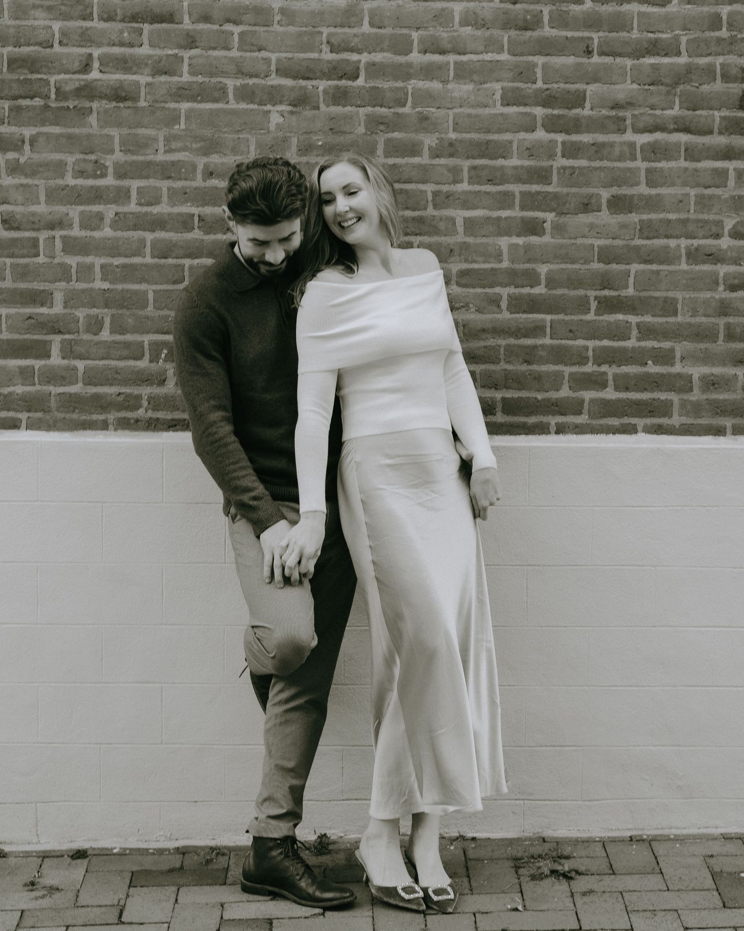 A couple in contrasting outfits poses intimately against a brick wall in a black and white photo.