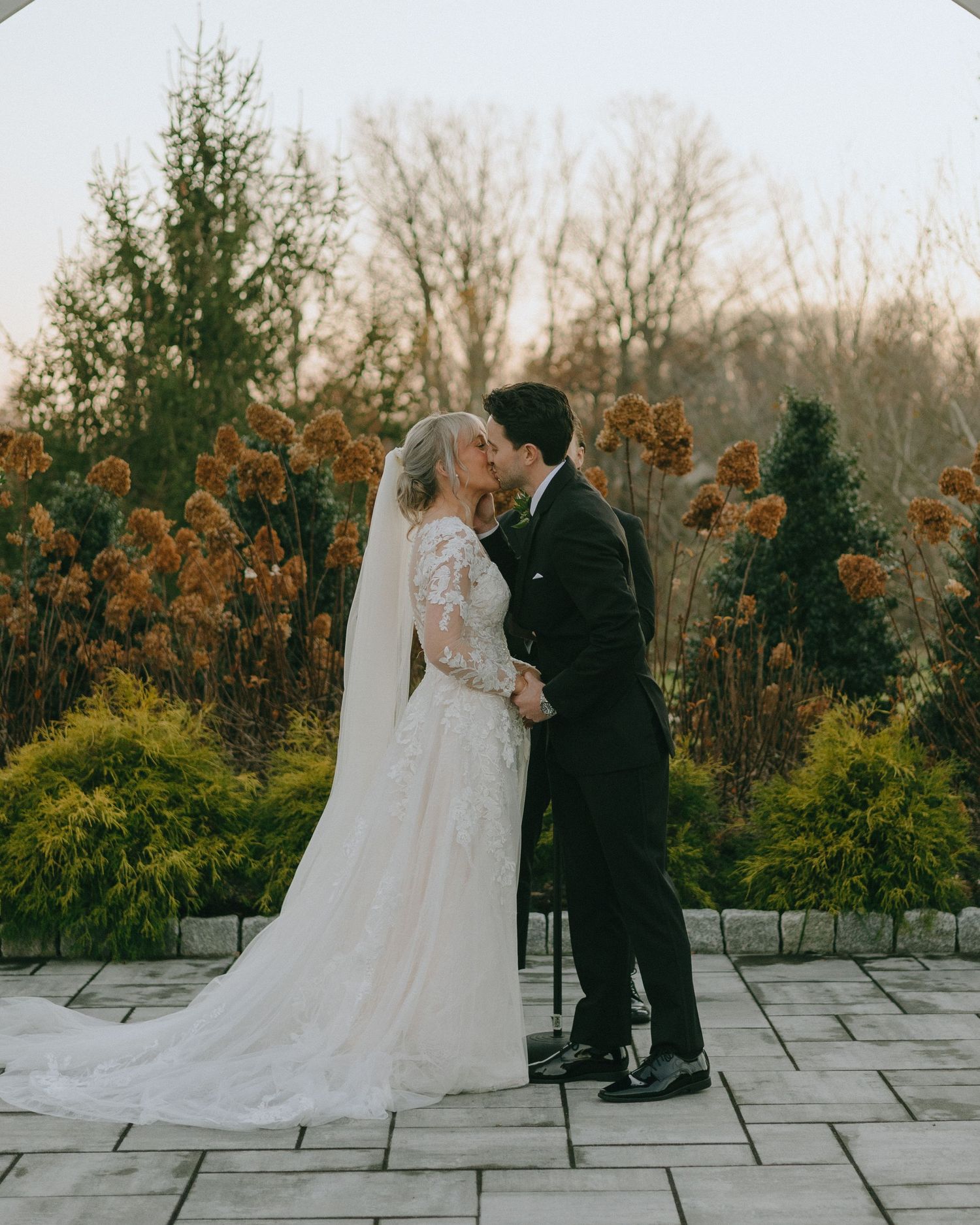A bride in a flowing white gown and groom in black suit share an intimate moment on a stone terrace.