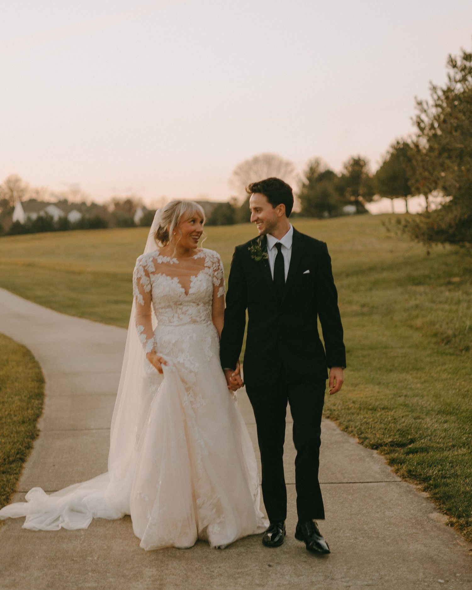 A couple in wedding attire walks together on a paved path during golden hour in a serene outdoor setting.