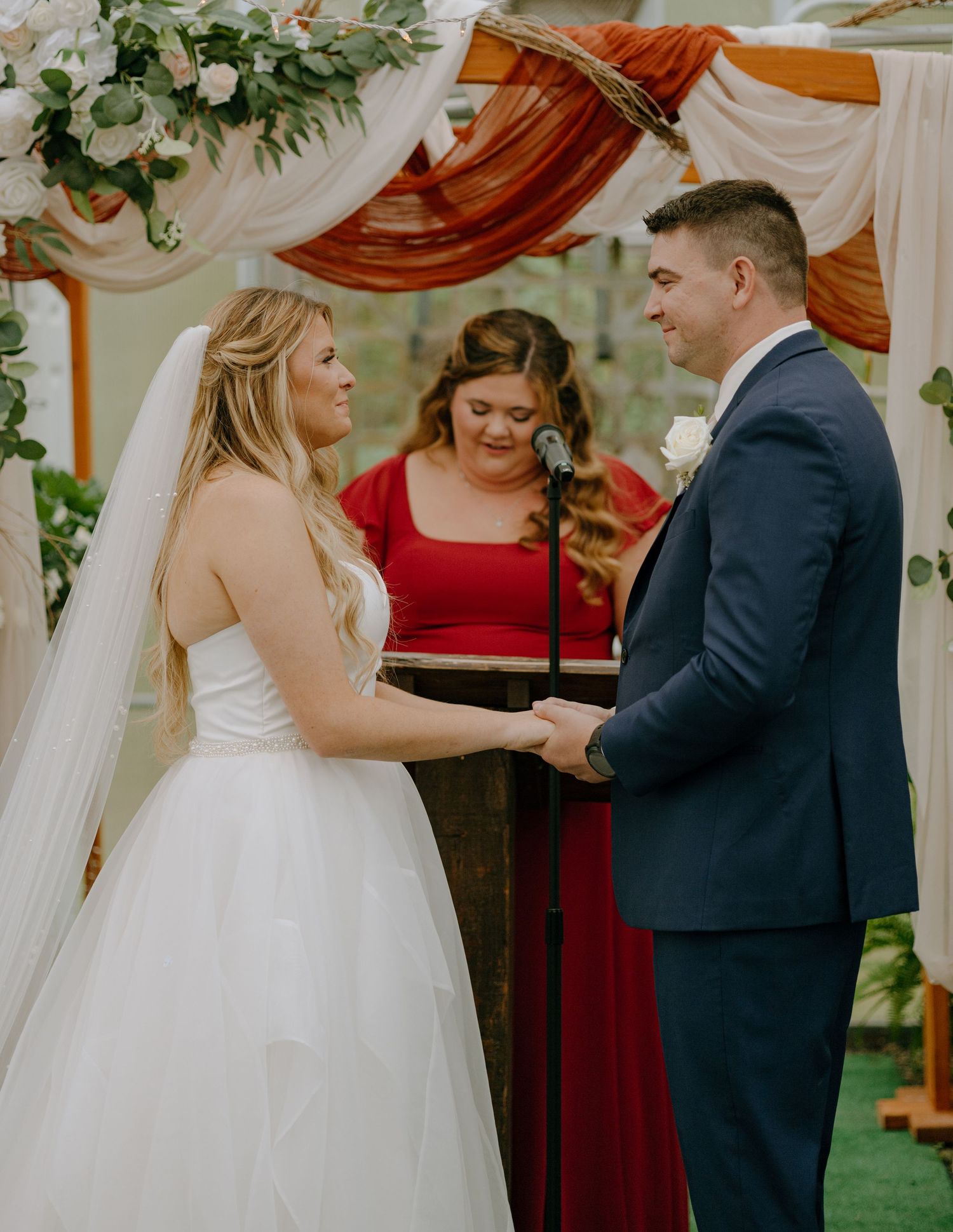 A wedding ceremony takes place under draped fabric decor with floral arrangements and attendants in burgundy dresses.