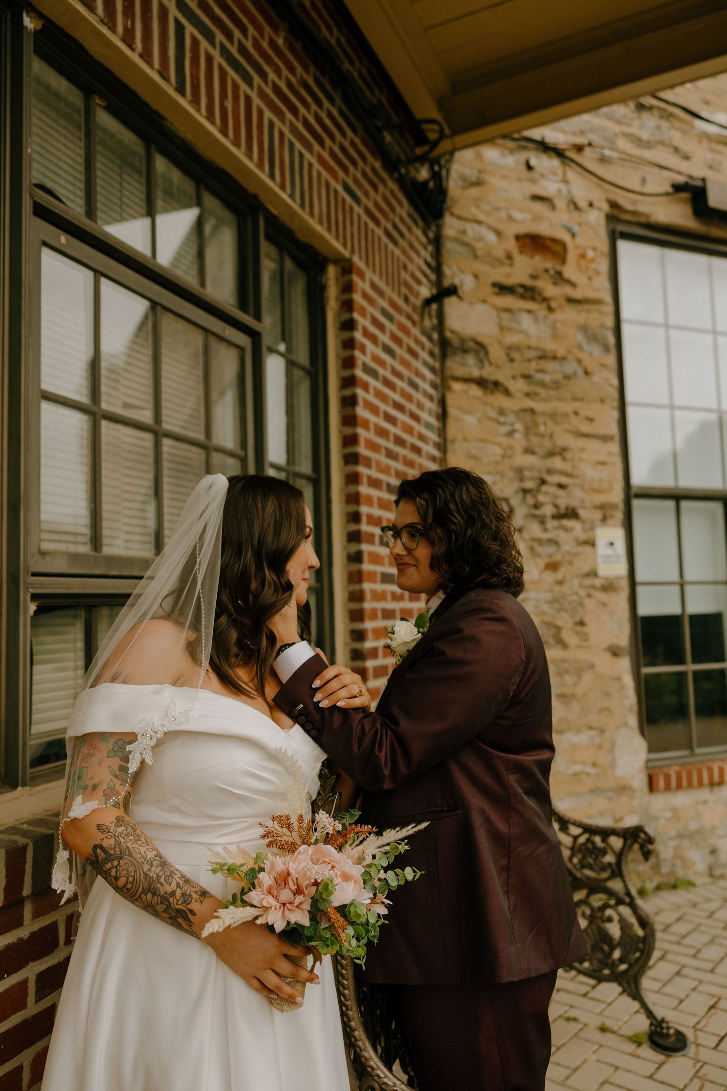 A romantic wedding couple embraces against a rustic brick and stone building exterior.