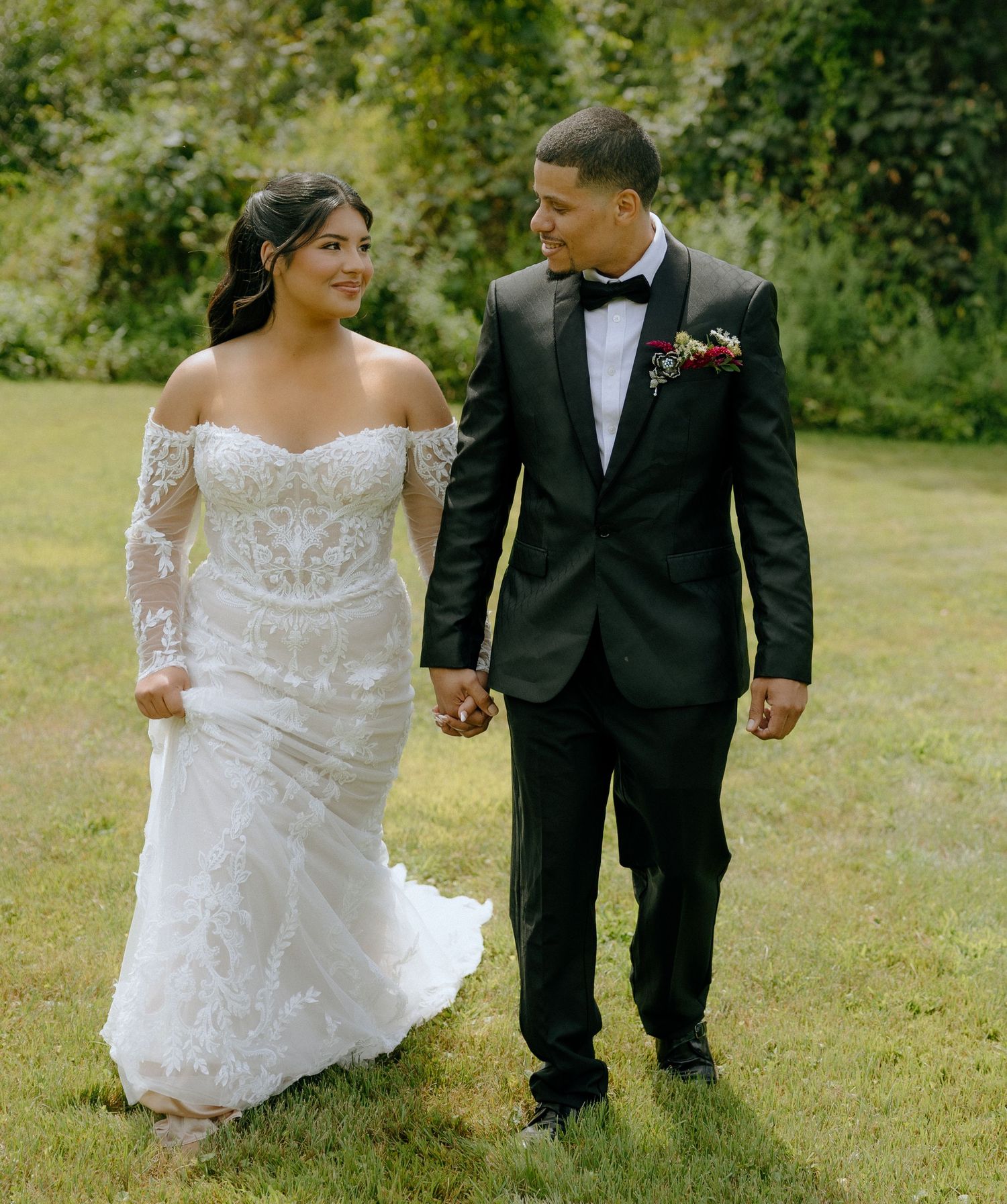 A couple in wedding attire walks together on a grassy lawn, with the bride wearing a lace gown.