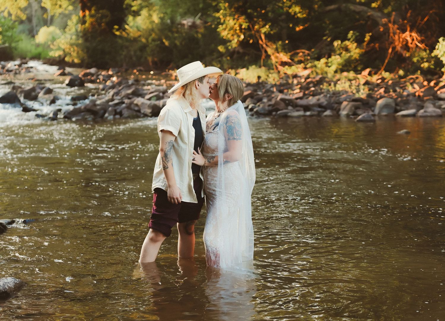 A couple shares an intimate moment while standing in a river with golden autumn foliage in the background.