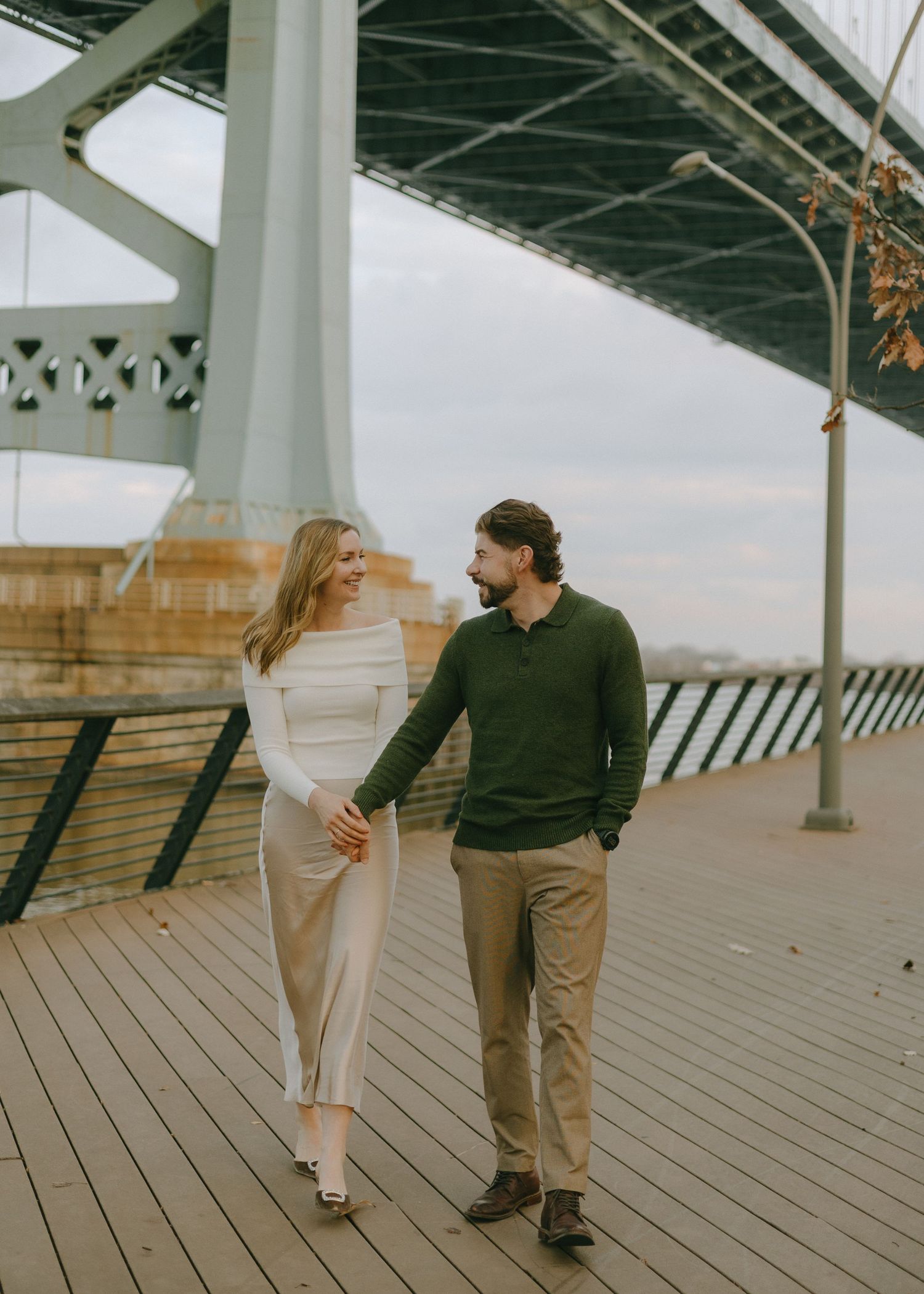 A couple takes a romantic stroll along a wooden boardwalk beneath a large bridge structure.