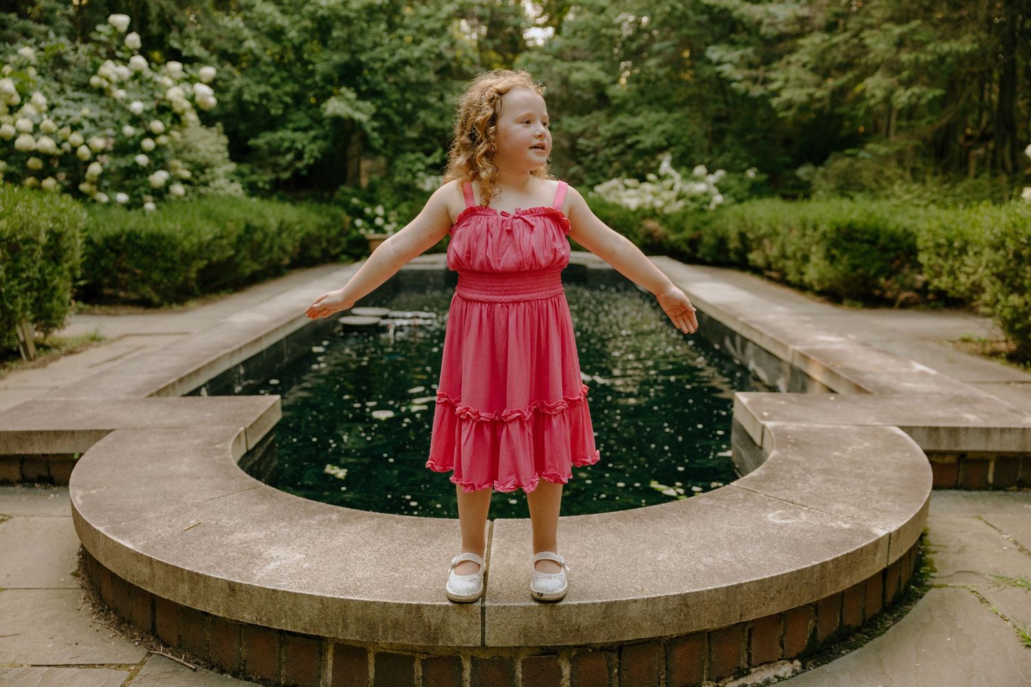 Two people in pink dresses play around circular stone feature in garden setting.