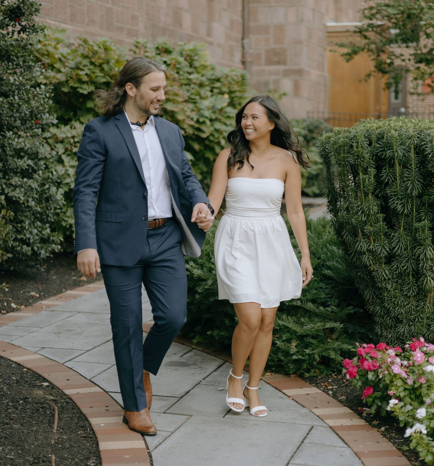 A couple shares a joyful moment together while walking along a stone pathway surrounded by lush gardens and flowers.
