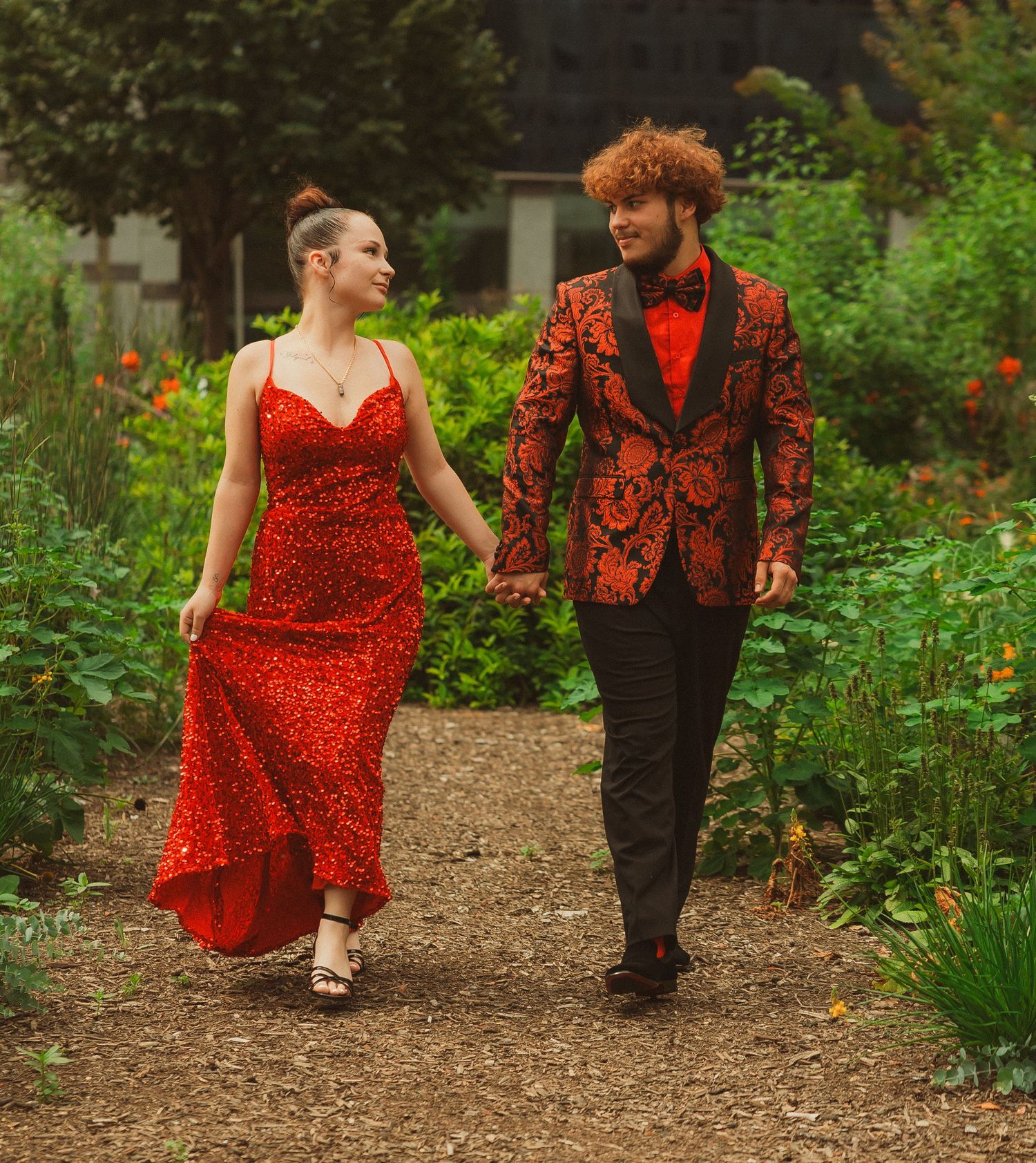 A couple in red and black outfits walks along a garden path surrounded by flowers.