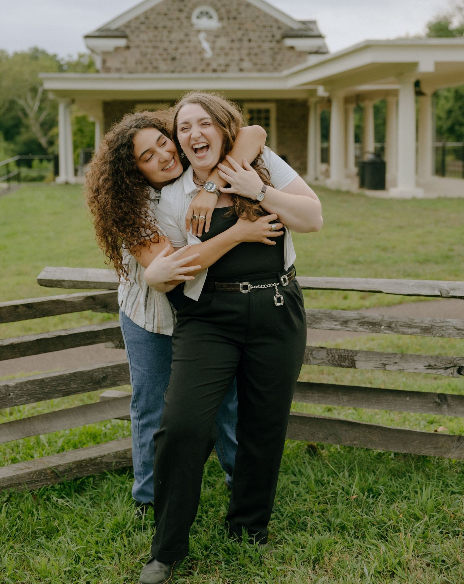 Two friends embrace and laugh together by a wooden fence with a building in background.