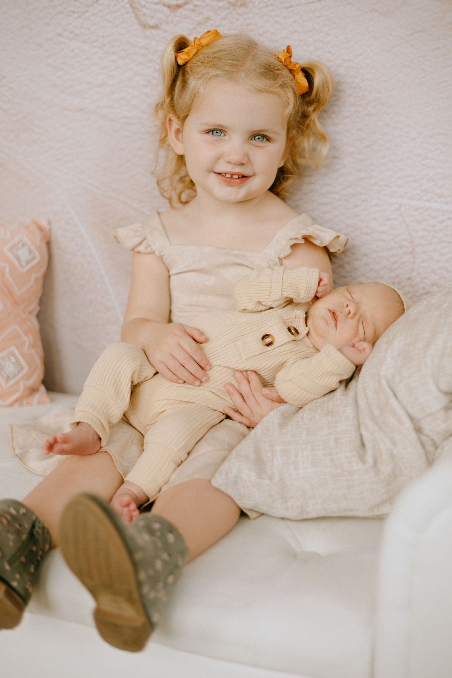 A young child in a cream dress relaxes on white bedding with vintage-style accessories.