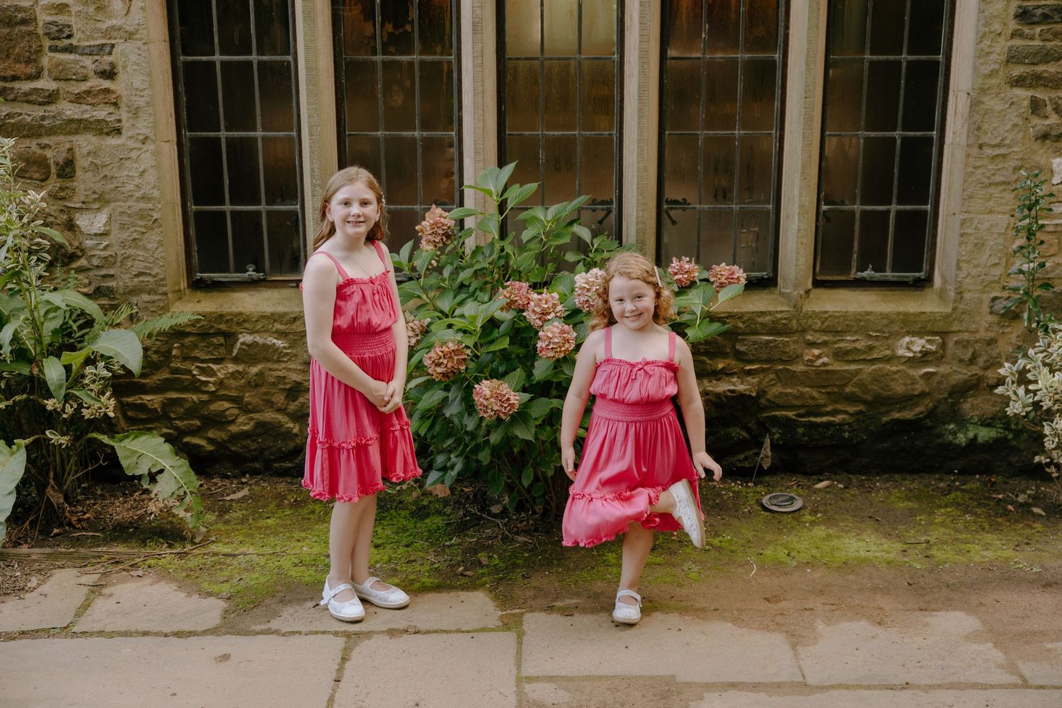 Two people in ruffled pink dresses stand against a stone building with windows.