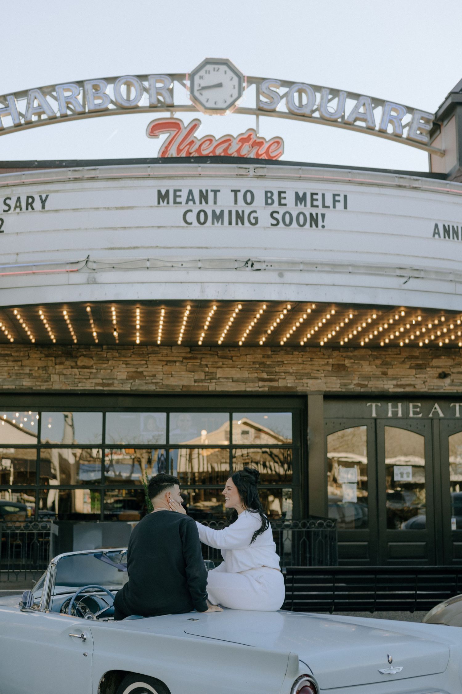 Classic cars parked outside a vintage movie theater with illuminated marquee signs.