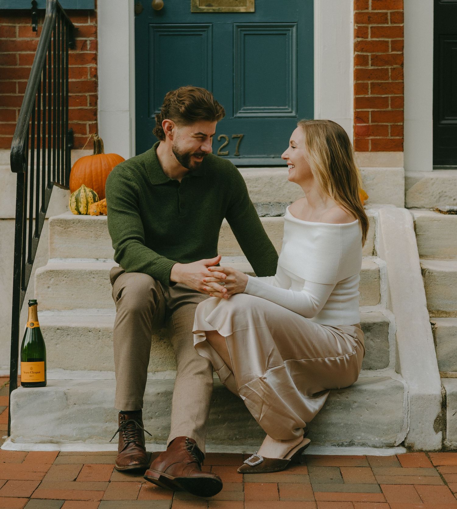 A couple shares an intimate moment sitting on brick steps with a champagne bottle and pumpkin nearby.