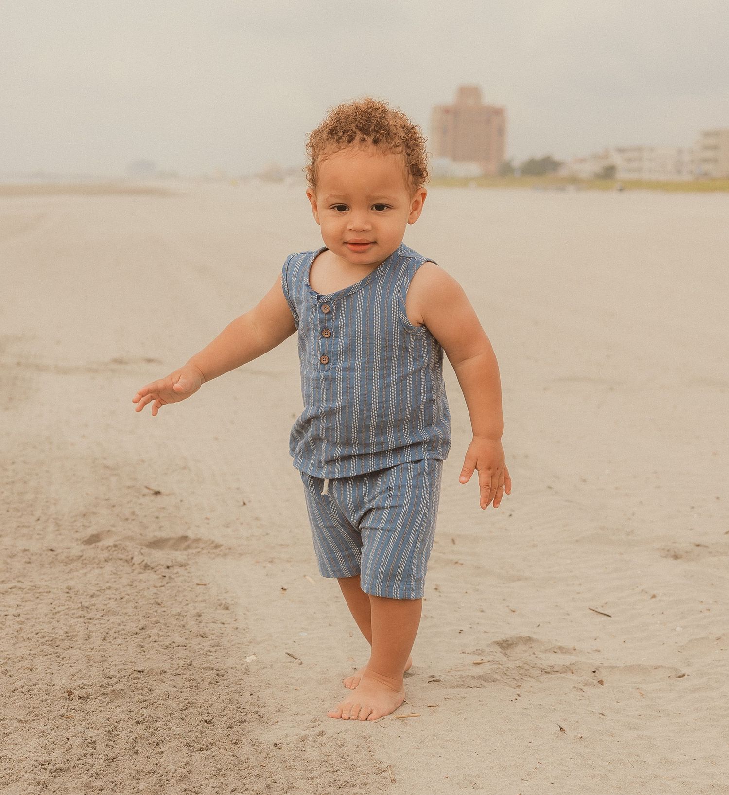 Toddler in blue striped romper exploring sandy beach barefoot.