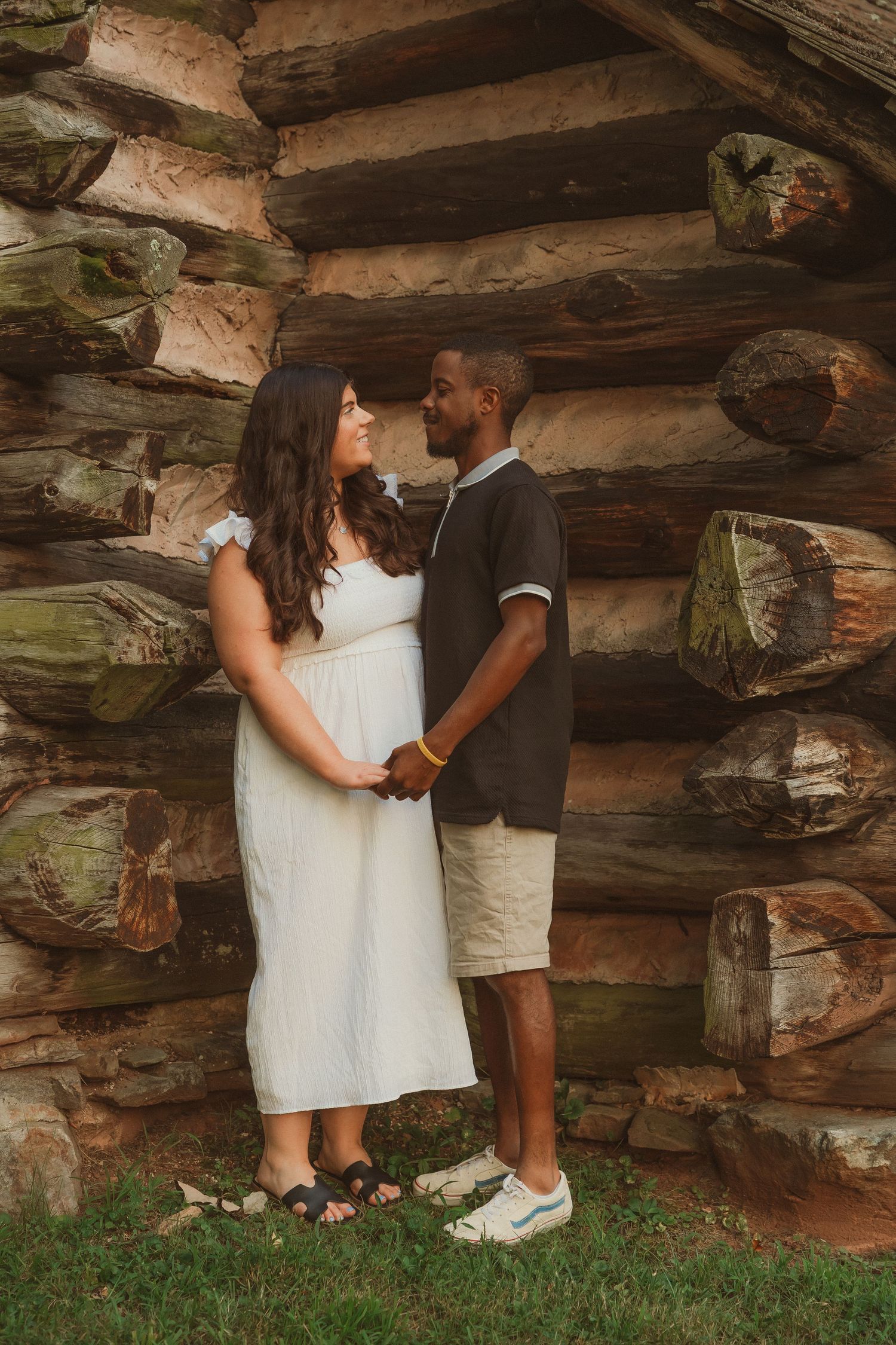 A couple poses against a rustic log cabin wall wearing casual attire.