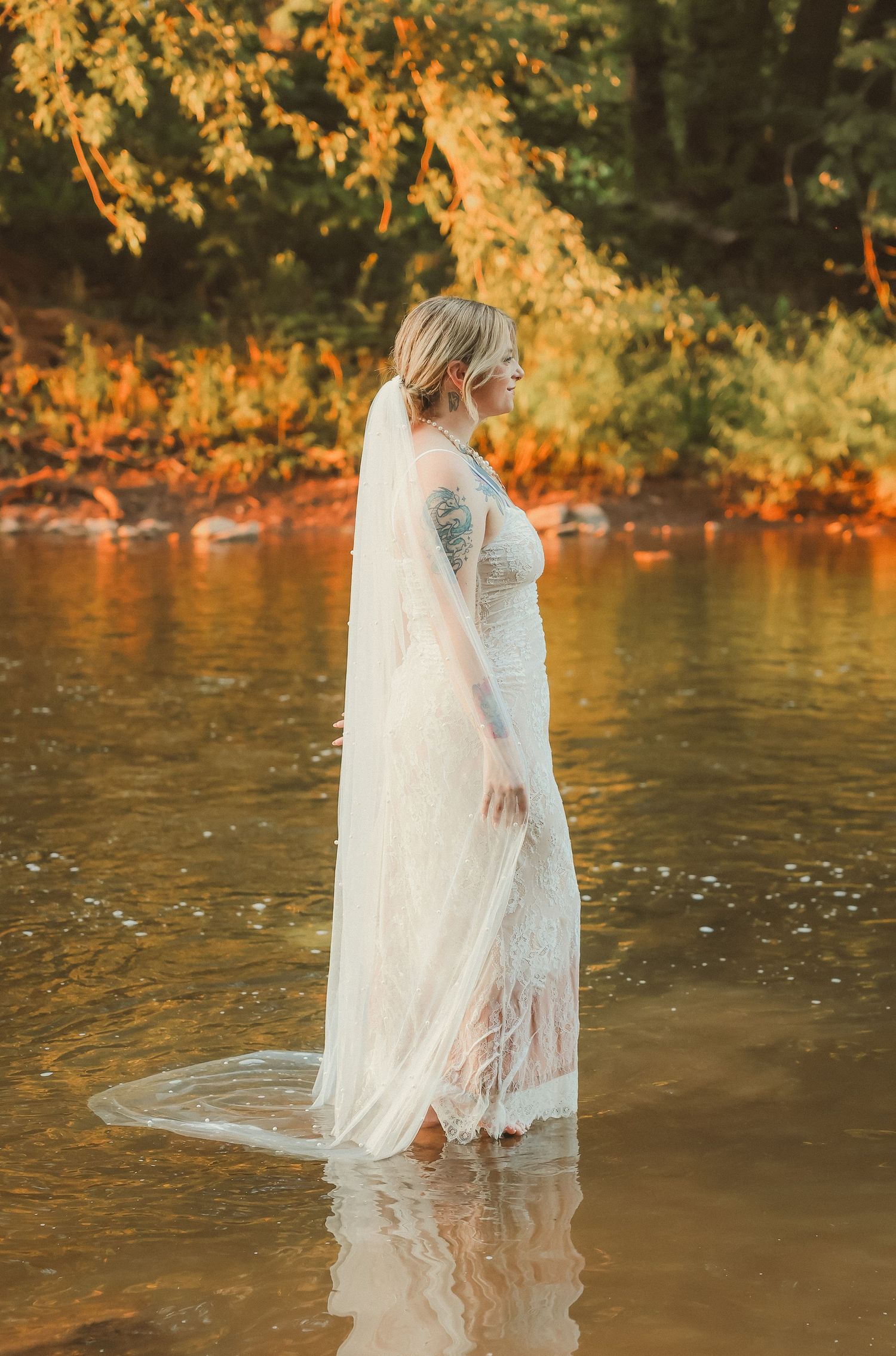 A person in a wedding dress stands in shallow river water during sunset.