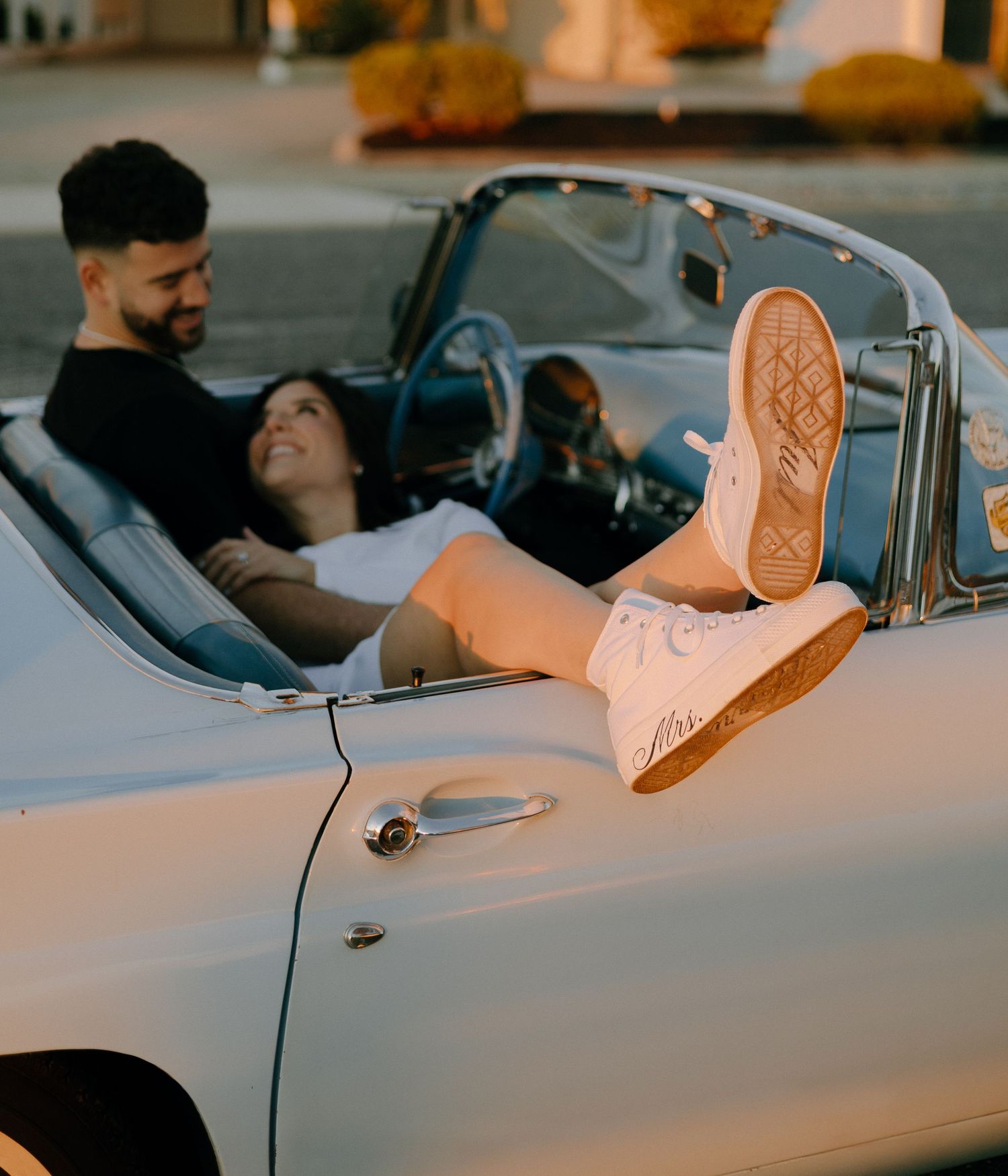 A playful couple relaxes in a classic convertible car with pink sneakers visible.