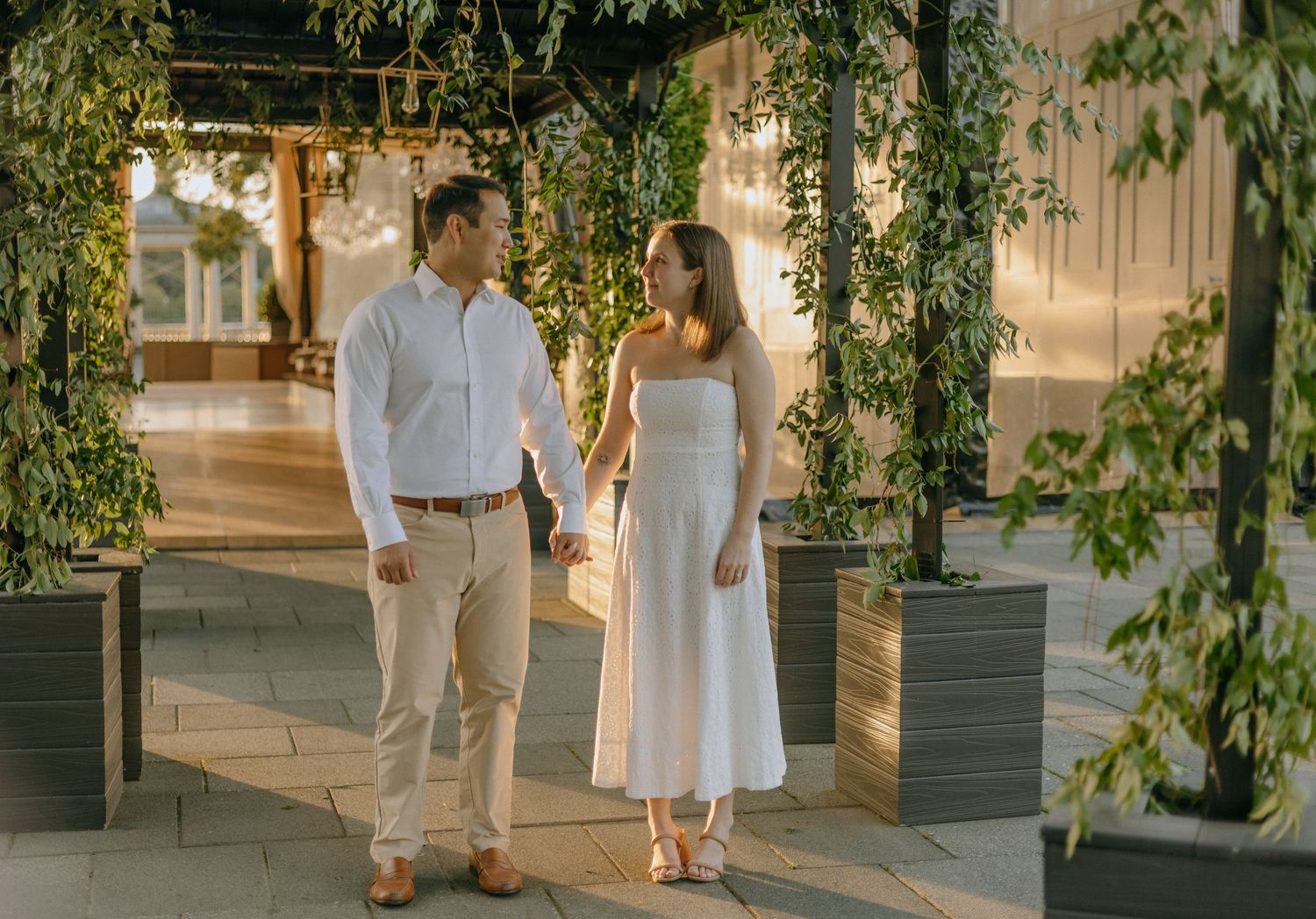 A couple in white outfits walks together under a pergola with hanging vines during golden hour.