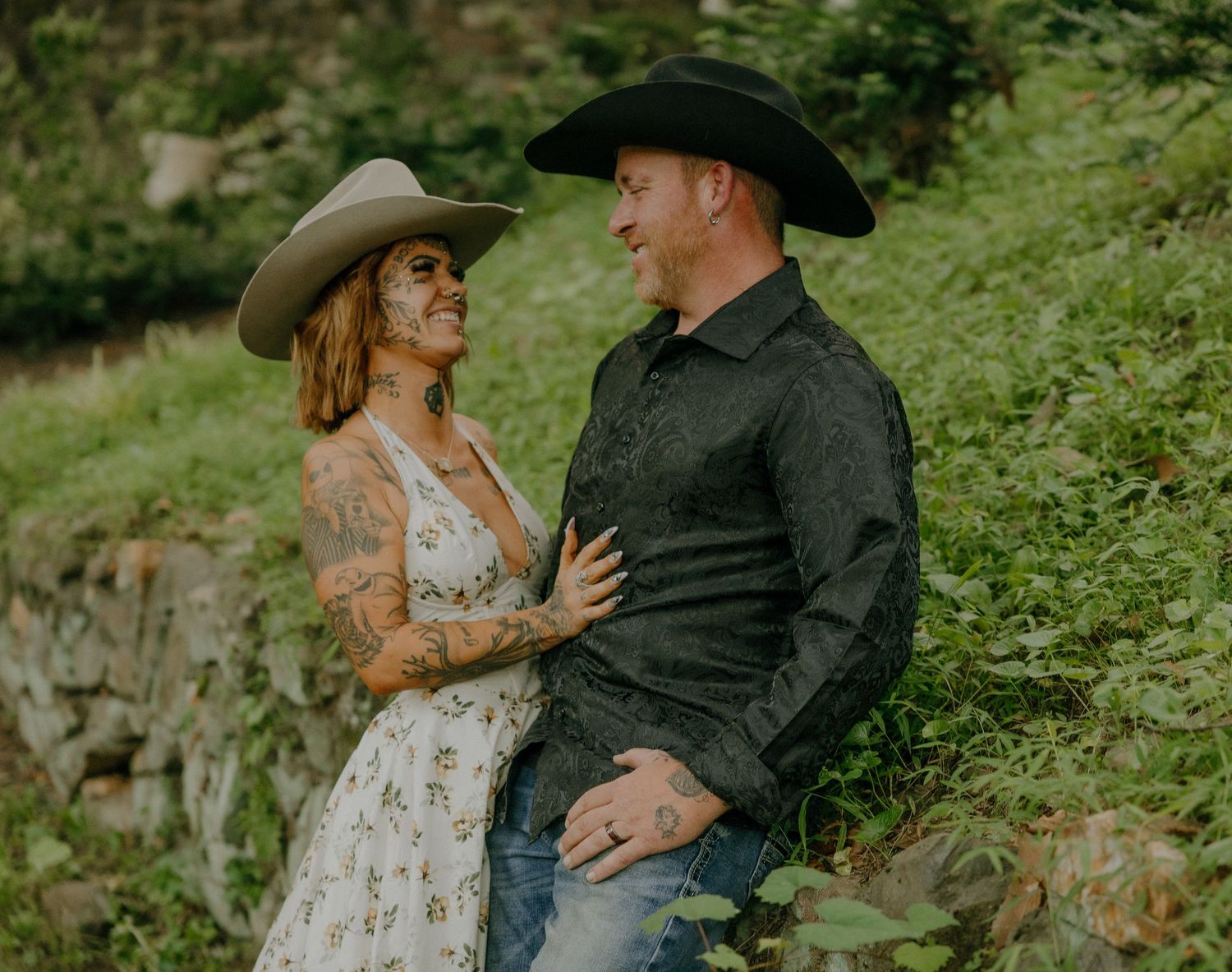 Couple in western wear share romantic moments against lush green backdrop.