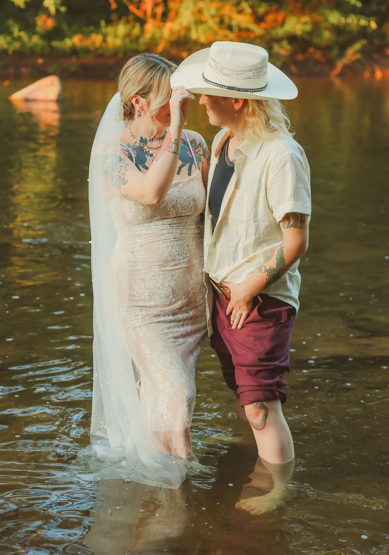 Two people wade through shallow river water during a sunset wedding photoshoot.