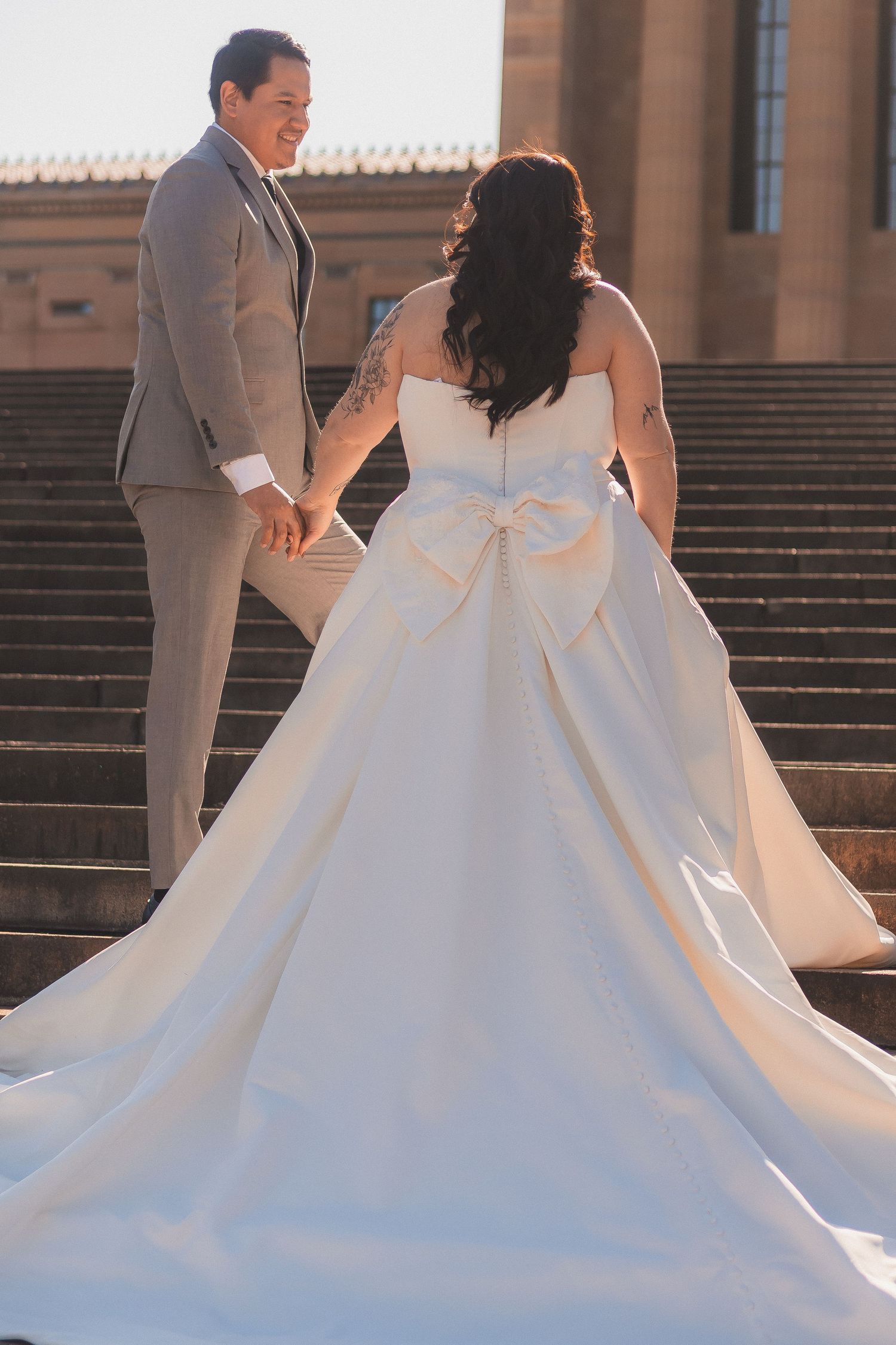 A flowing white wedding dress with a dramatic bow detail captured from behind on stone steps at sunset.