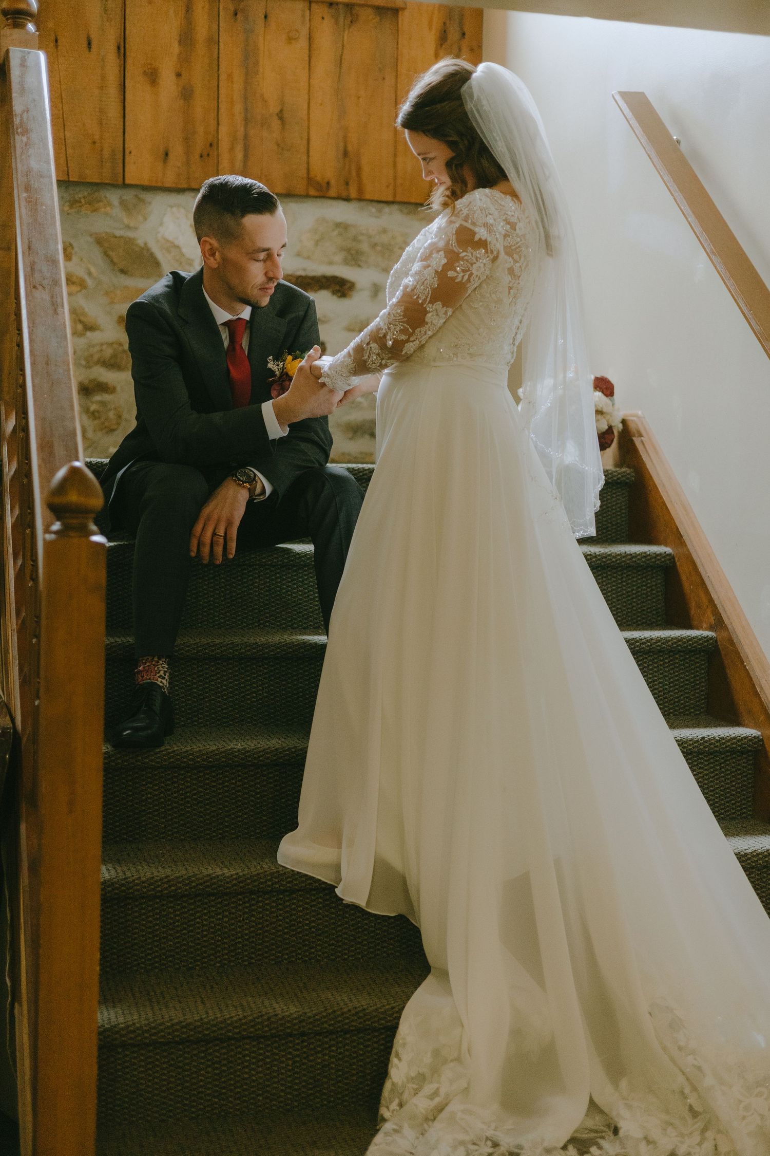 Bride in flowing white wedding dress sits on wooden staircase in rustic venue during intimate moment.