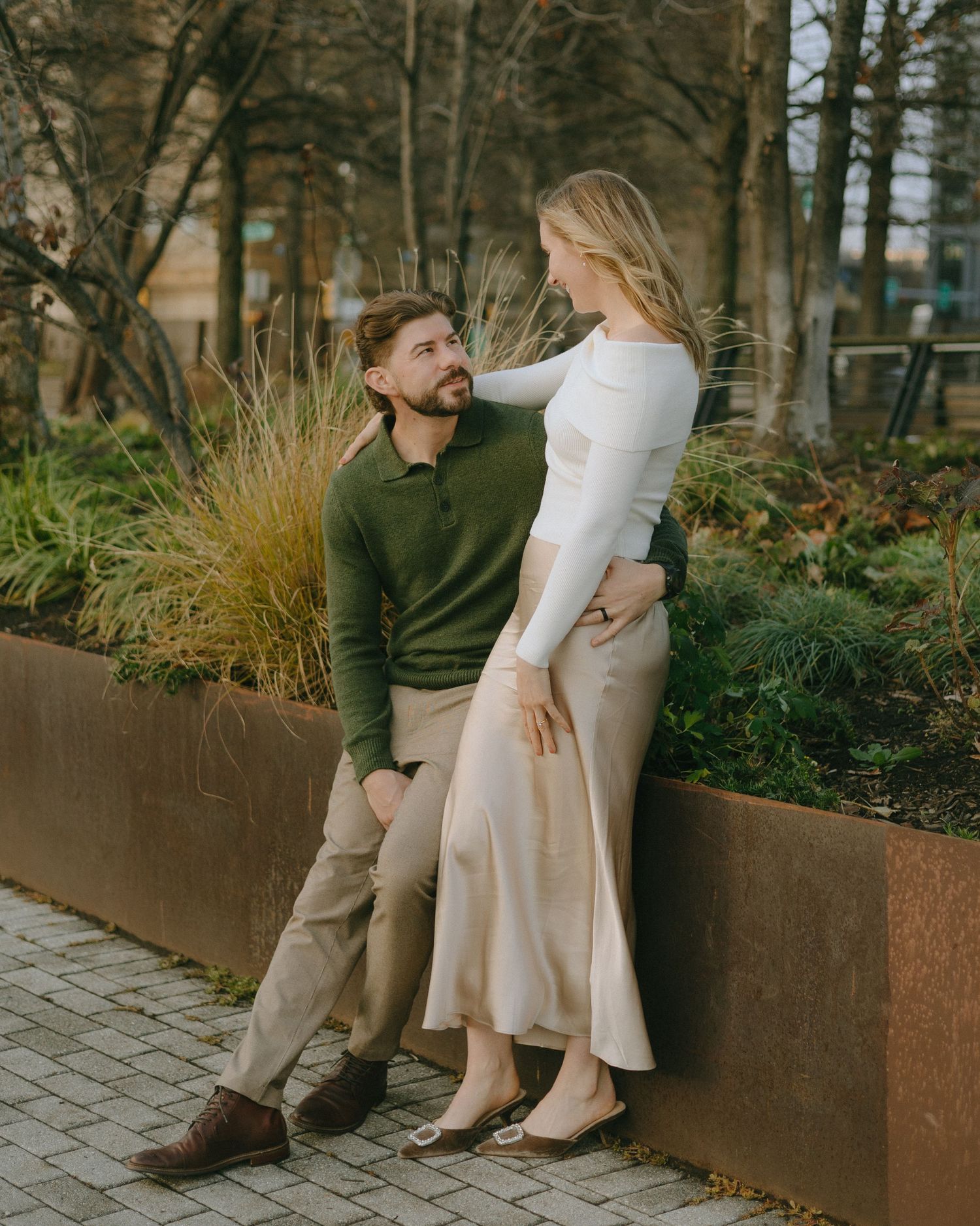 A couple sits intimately on a concrete planter surrounded by autumn foliage in an urban setting.
