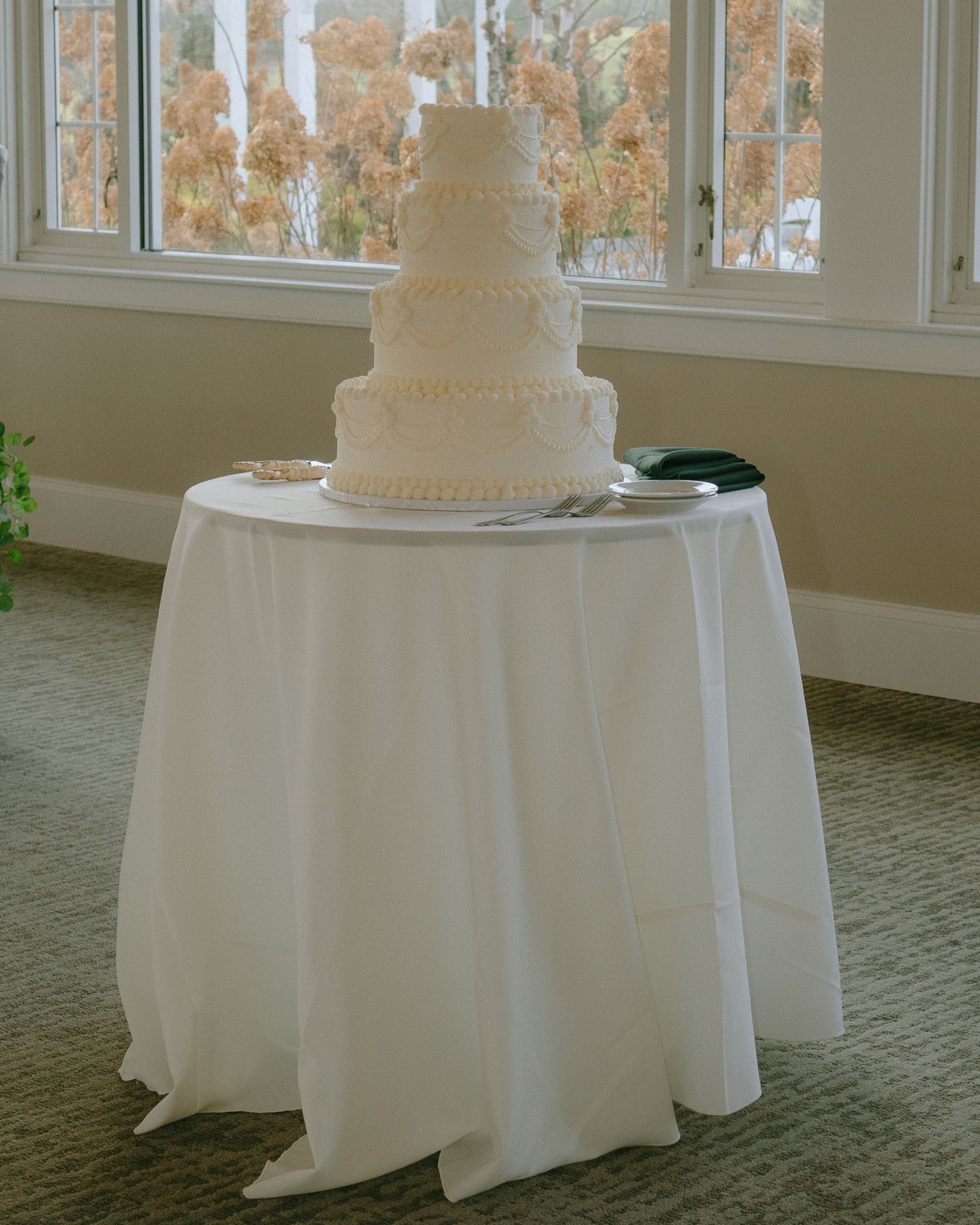 A classic white tiered wedding cake sits on a round table with white tablecloth near windows overlooking autumn trees.