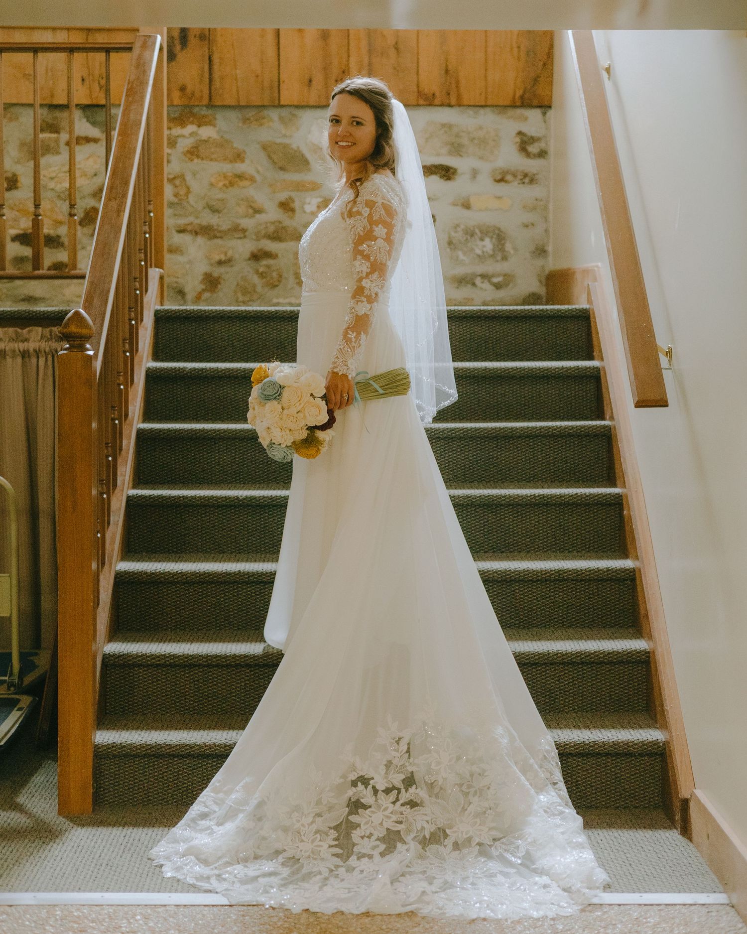 A bride in a flowing white wedding gown with long train stands elegantly on rustic wooden stairs in a vintage setting.