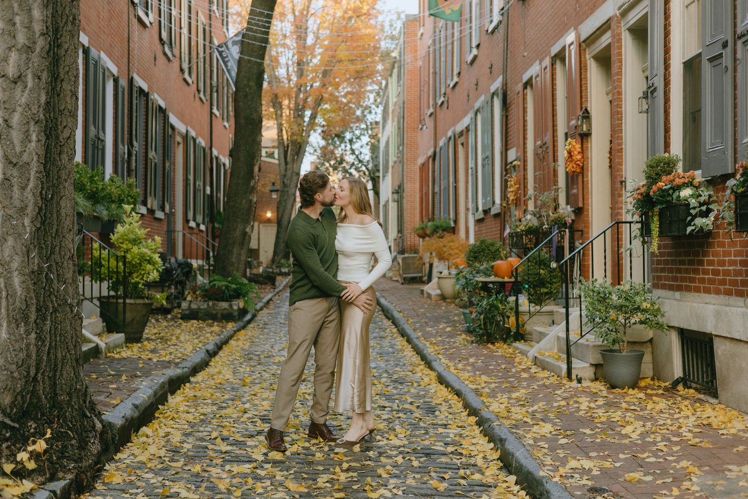 A couple stands together on a charming cobblestone street lined with autumn leaves and historic brick buildings.